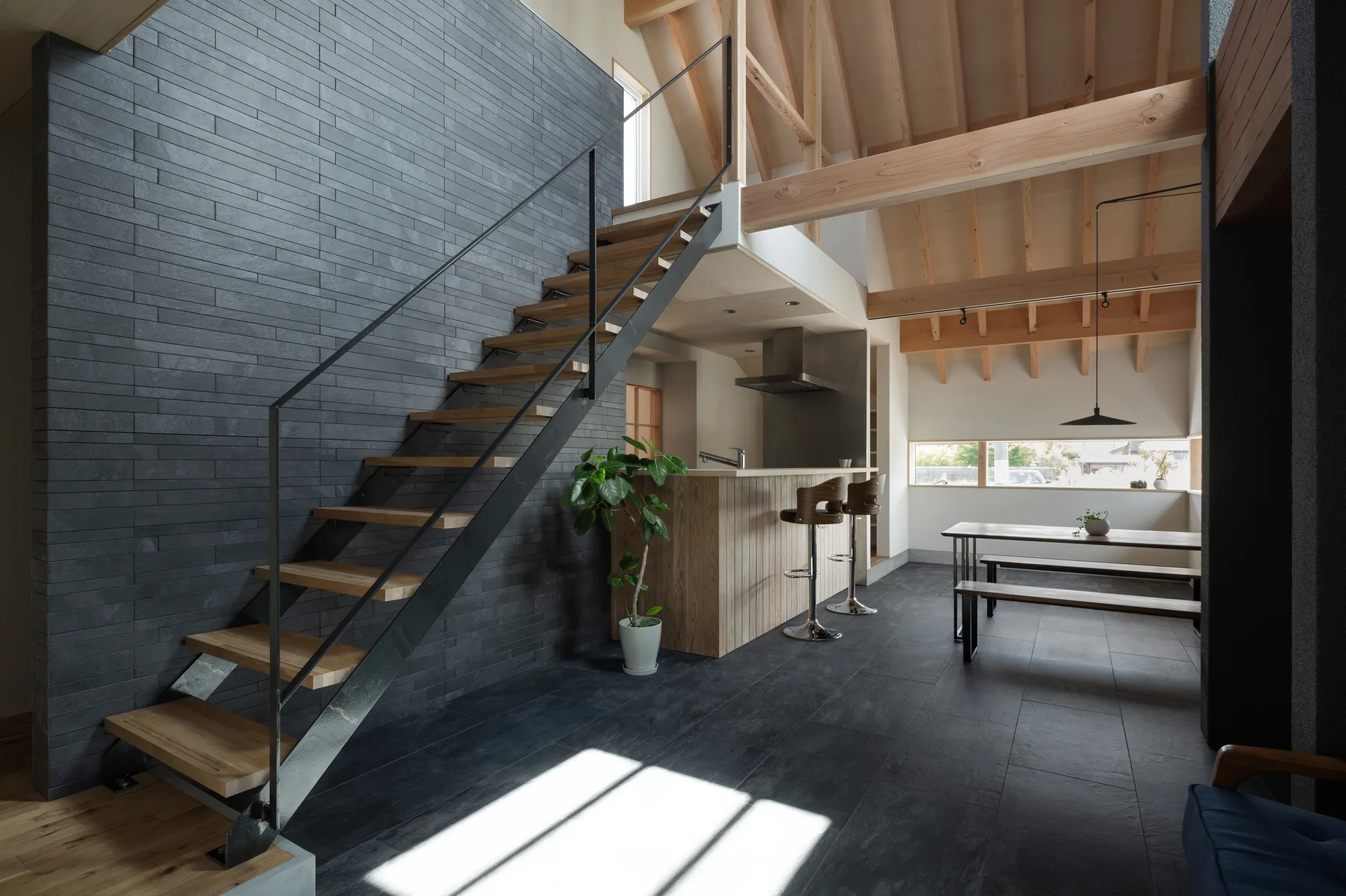 A modern open-plan interior features a wooden staircase against a dark gray tiled wall, with an exposed wood ceiling and kitchen island.