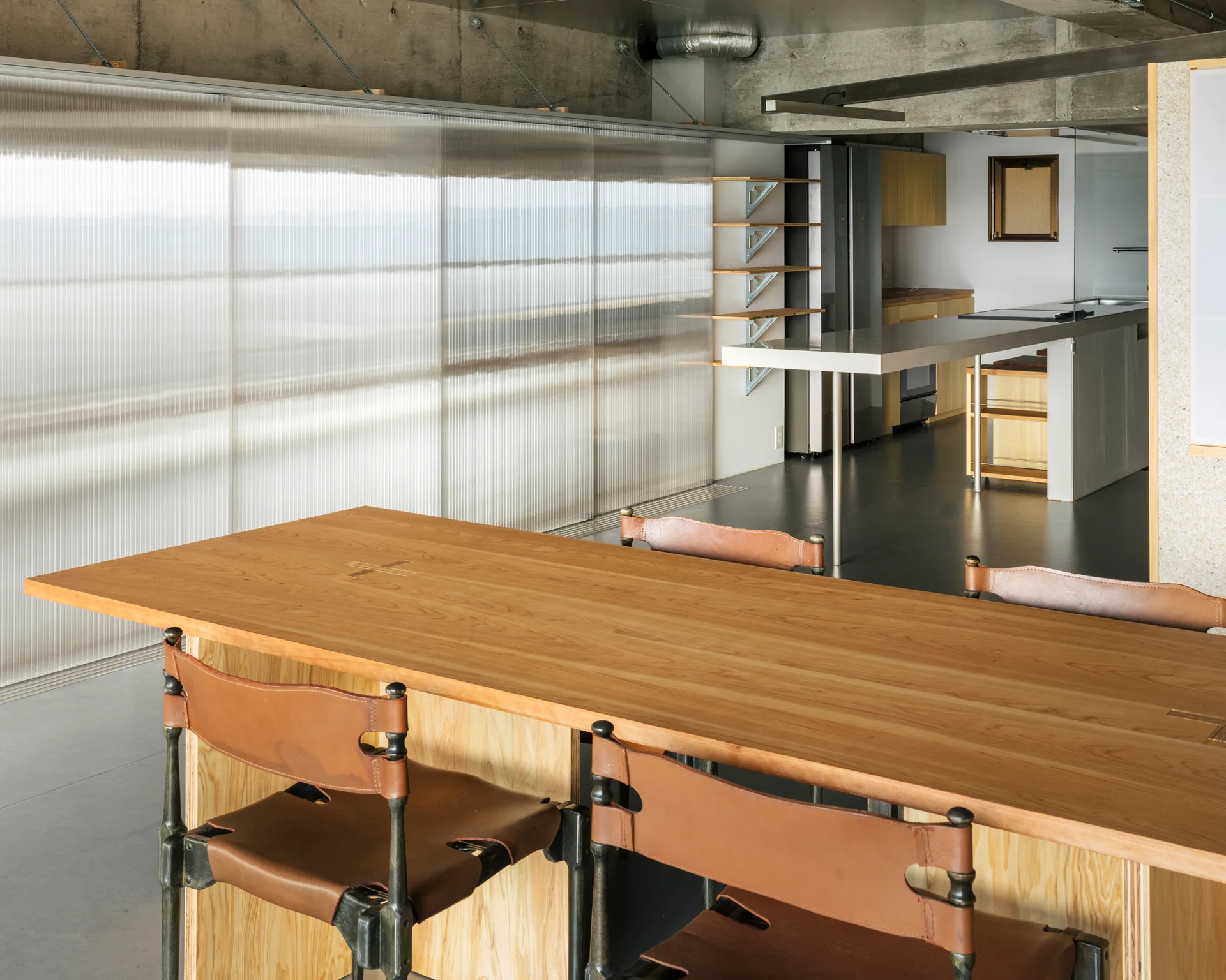 A wooden dining table with leather chairs sits before a large corrugated translucent sliding wall in an industrial space with an exposed concrete ceiling.