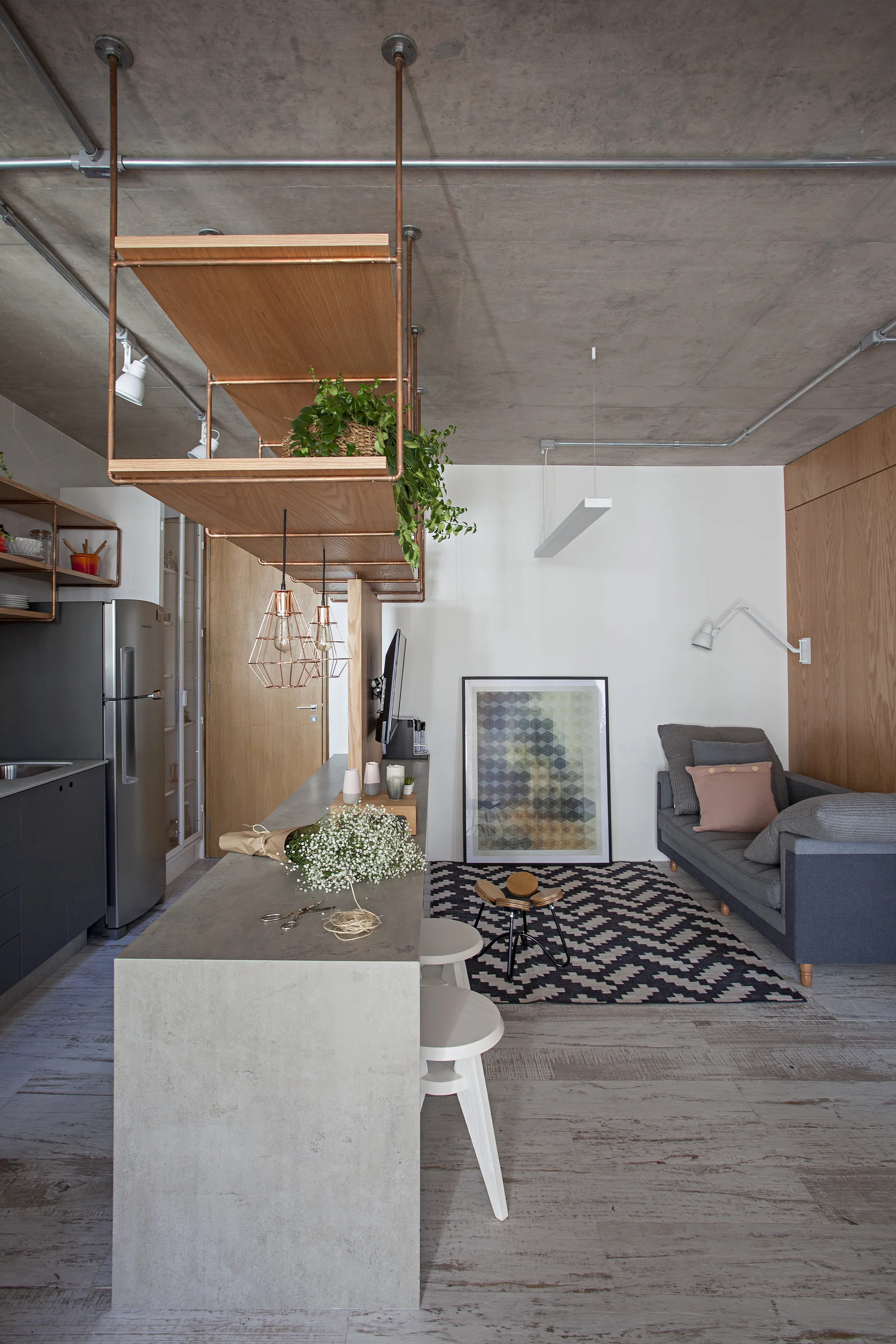An open-concept apartment featuring a concrete island, grey sofa, geometric rug, and suspended copper shelving beneath an exposed concrete ceiling.