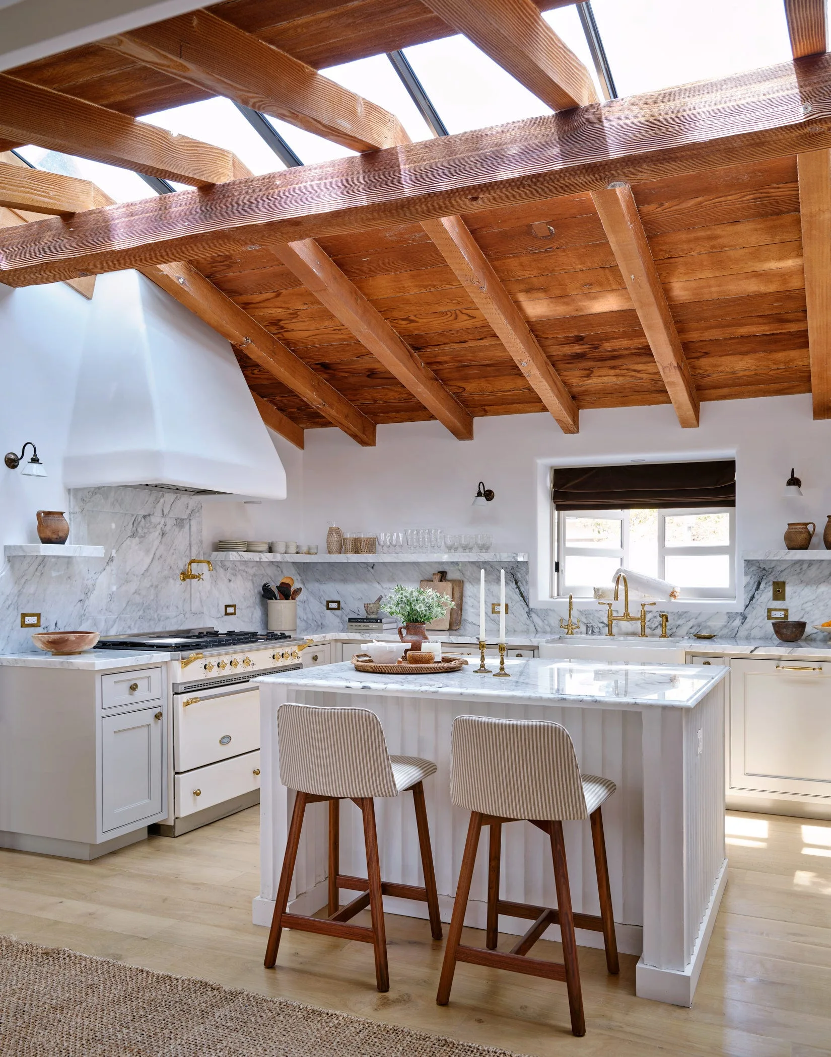 A bright kitchen with a marble island, striped barstools, white cabinetry, and a pitched ceiling with exposed wood beams and a skylight.