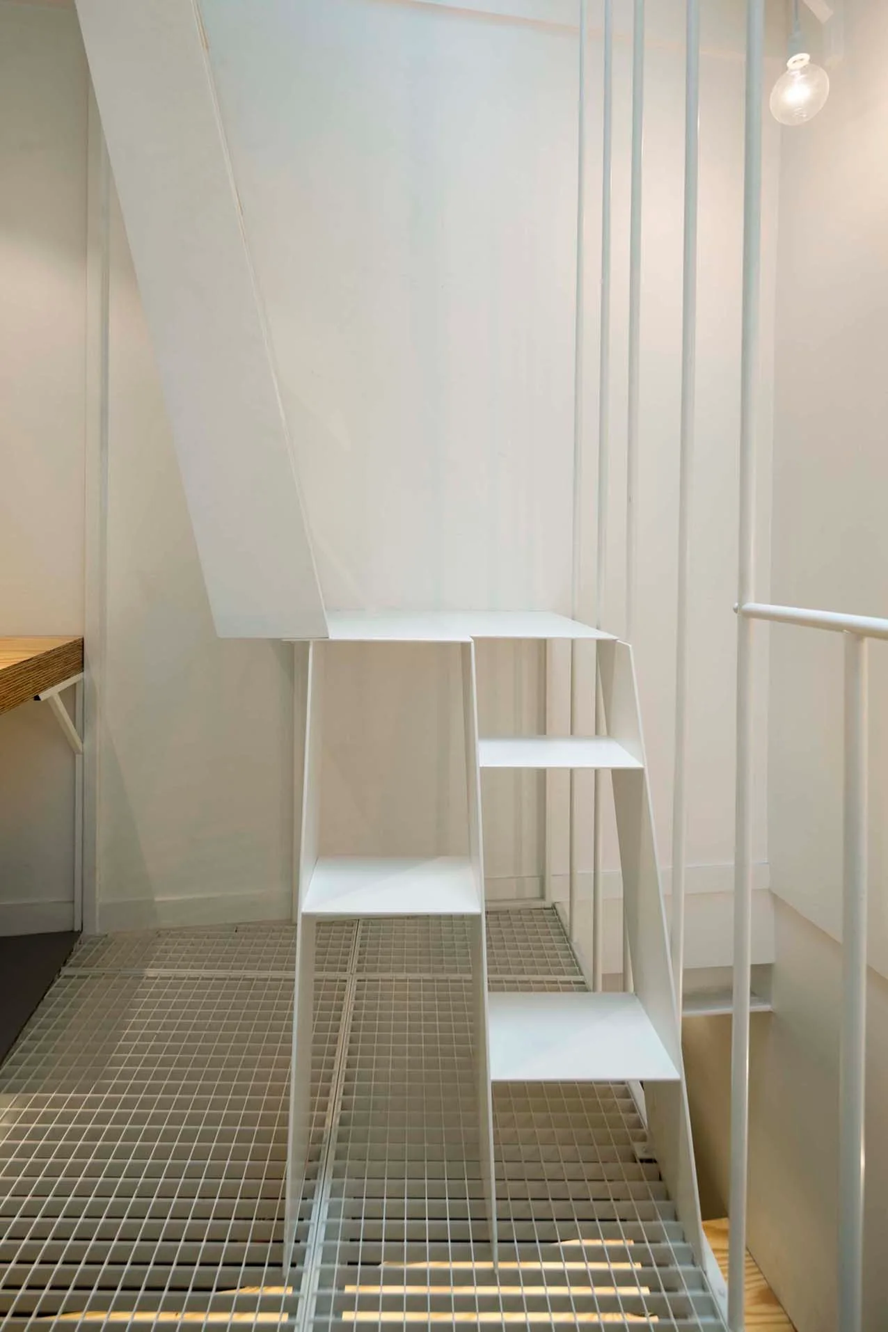 A minimalist white metal staircase with vertical railings ascends from a white gridded floor platform against a plain white wall.
