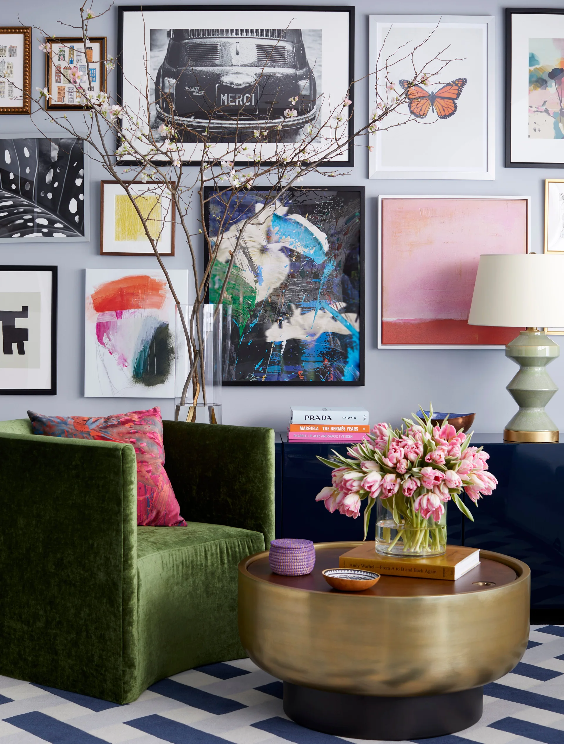 A green velvet armchair rests by a gold coffee table topped with pink tulips in front of a diverse gallery wall and blue console.