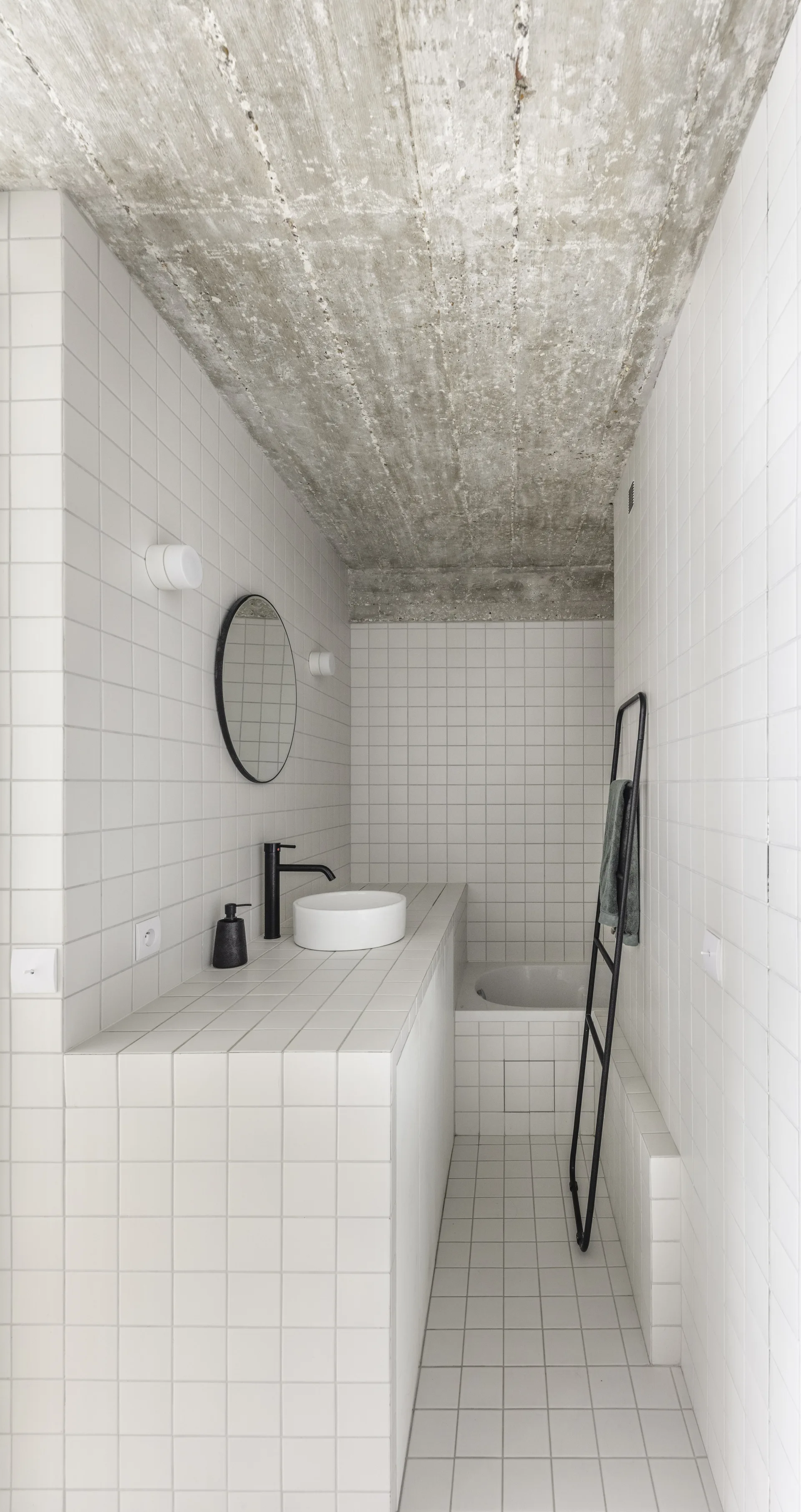 A minimalist bathroom featuring white square tiles on the floor and walls, an exposed concrete ceiling, a vessel sink, and a black ladder towel rack.