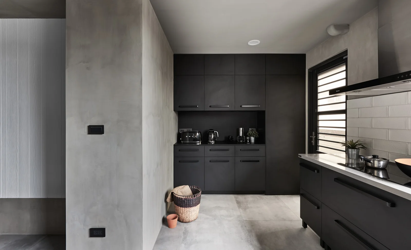 A minimalist kitchen featuring matte black cabinetry, grey concrete-textured walls, large floor tiles, and a white tiled backsplash.