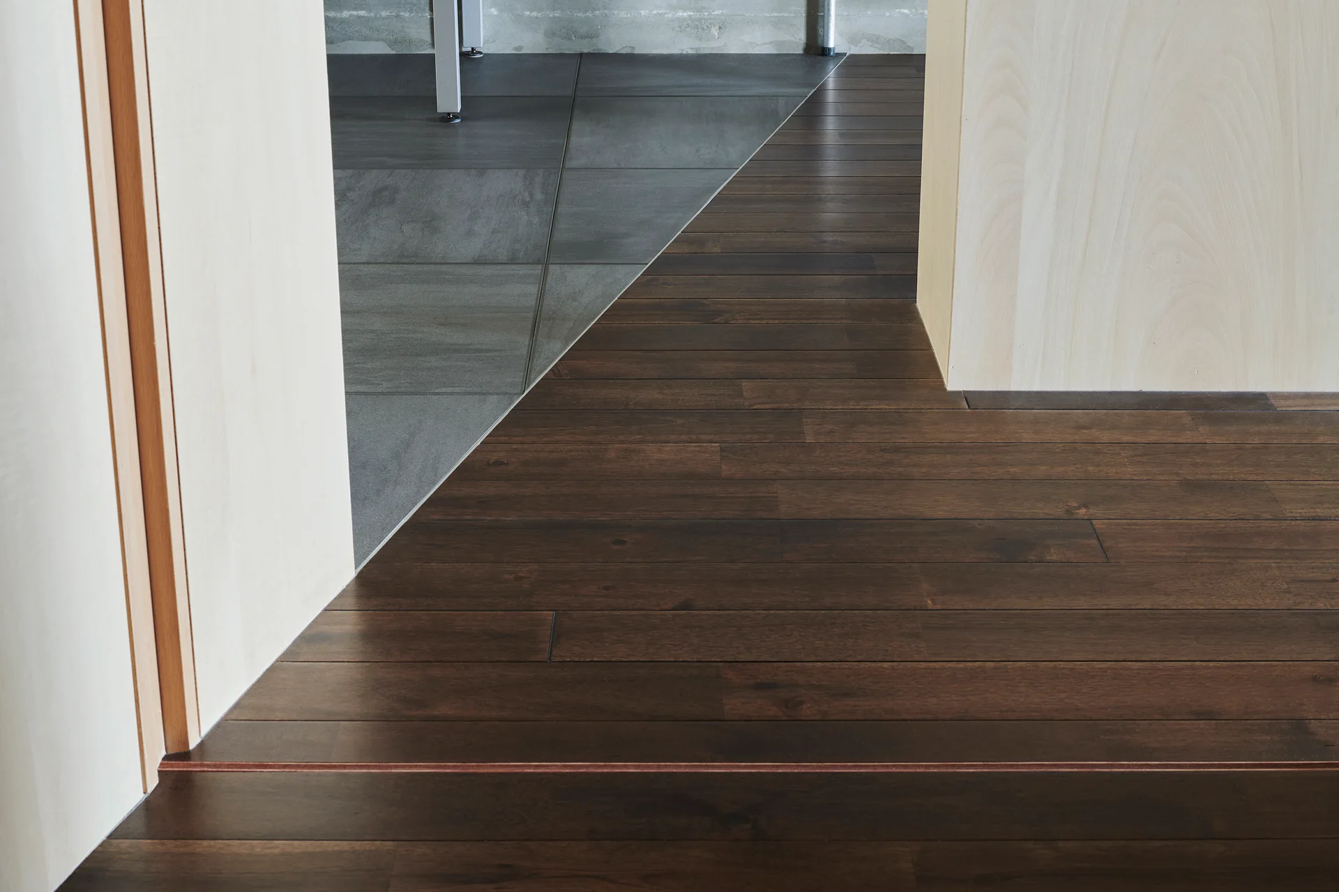 A close-up of a diagonal floor transition where dark brown wood planks meet large grey stone-like tiles next to light wood paneled walls.