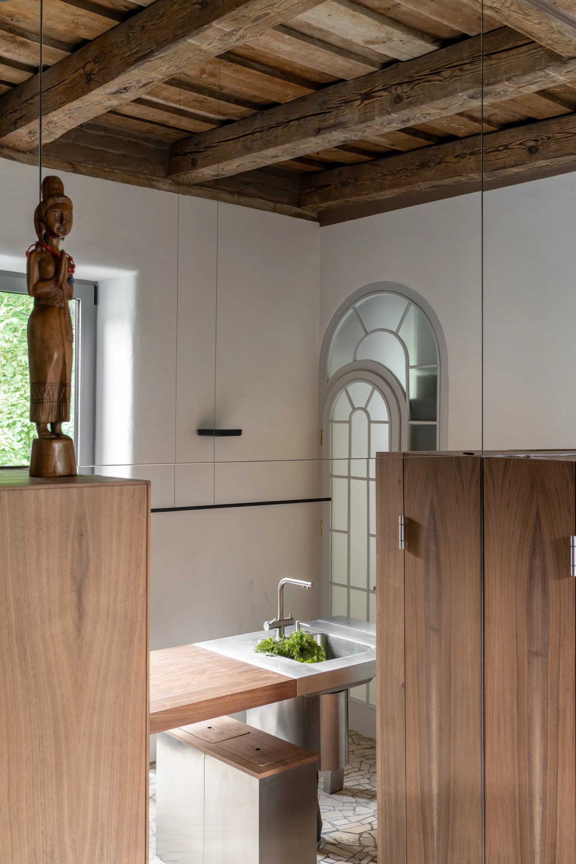 A modern kitchen featuring a stainless steel sink island, warm wooden cabinets, an arched glass door, and exposed rustic timber ceiling beams.