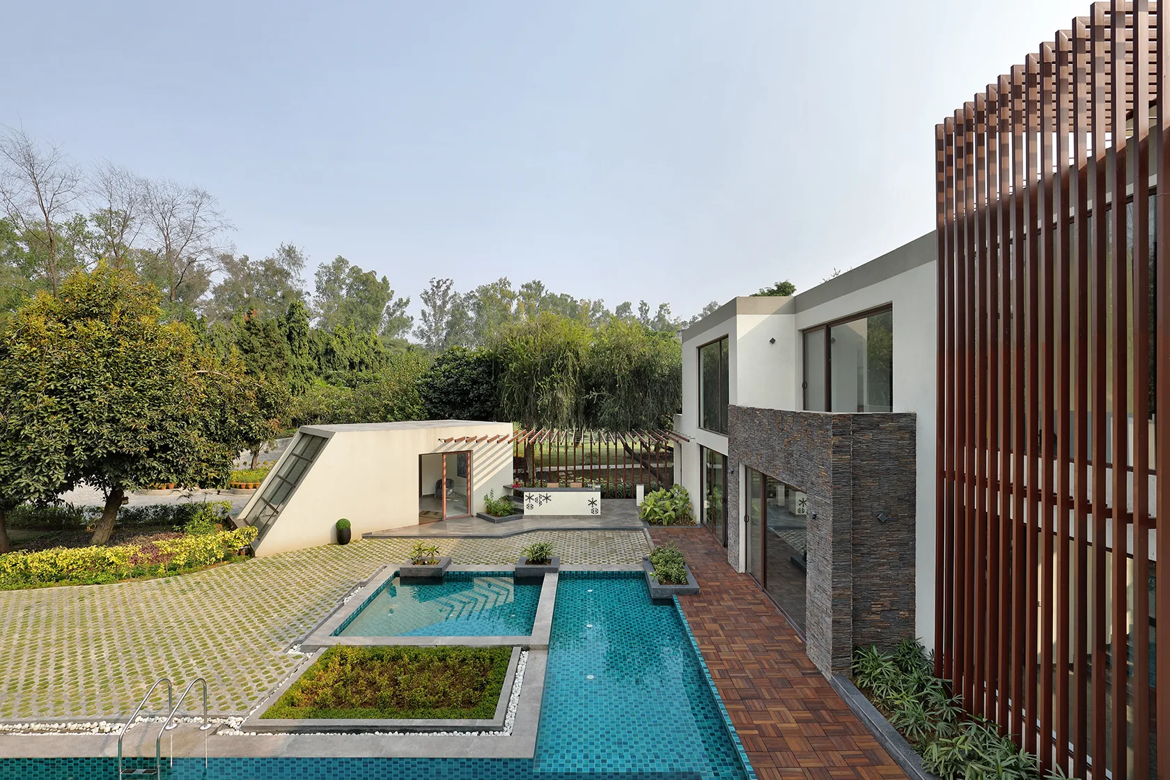 An aerial view of a modern backyard with a turquoise tiled pool, a wood deck, concrete grass pavers, and a contemporary house with a slatted screen.
