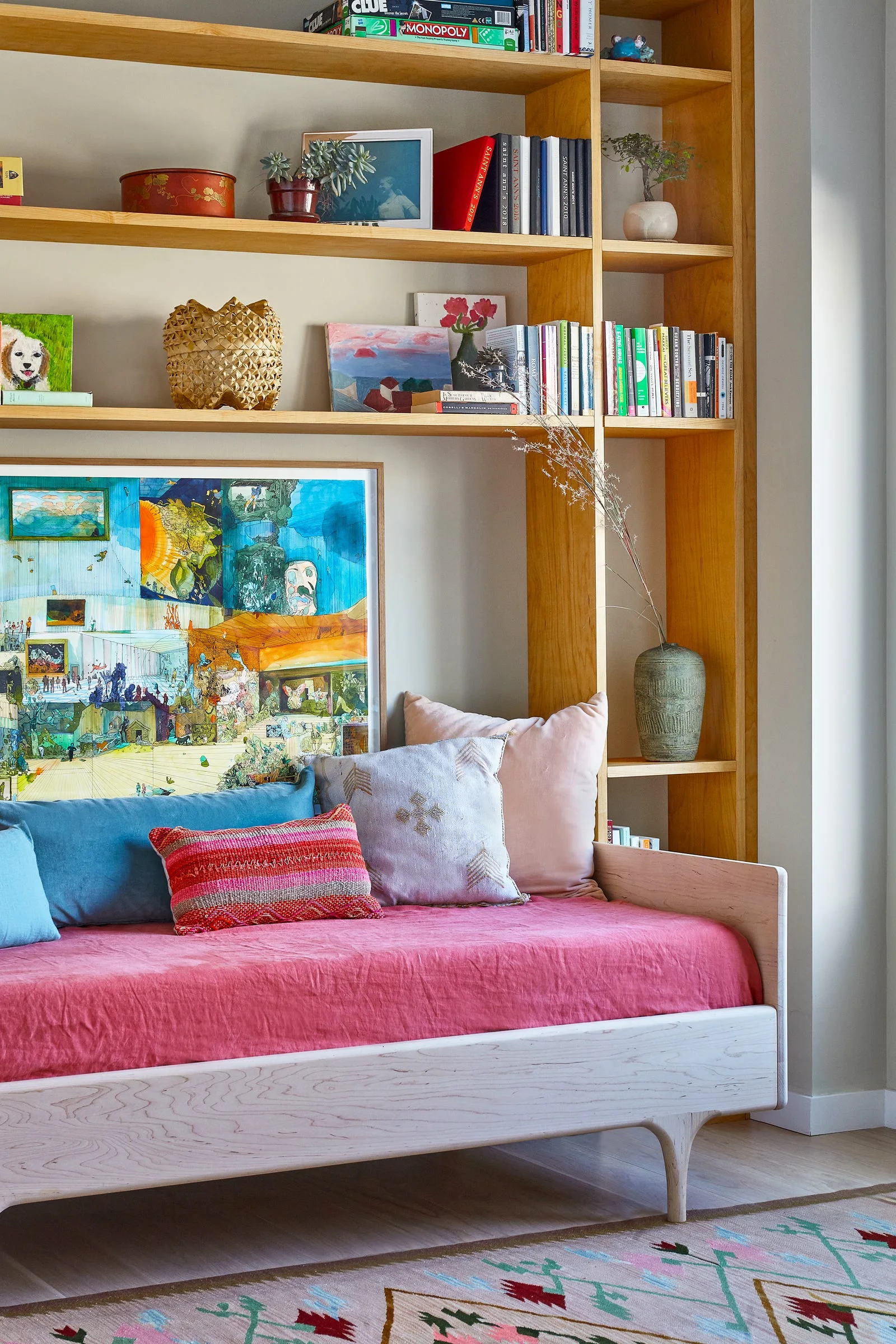 A light wood daybed with colorful pink and blue cushions sits nestled against a large built-in wooden bookcase displaying books and framed artwork.