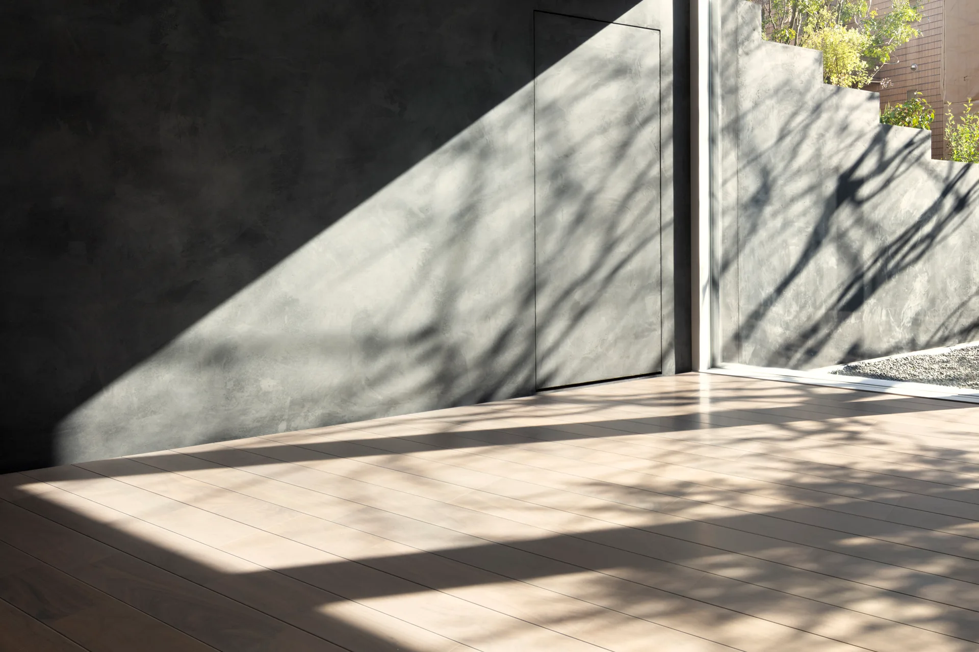 Natural sunlight casts tree shadows across a modern patio featuring light wood plank decking, a dark grey textured wall, and a seamless flush door.