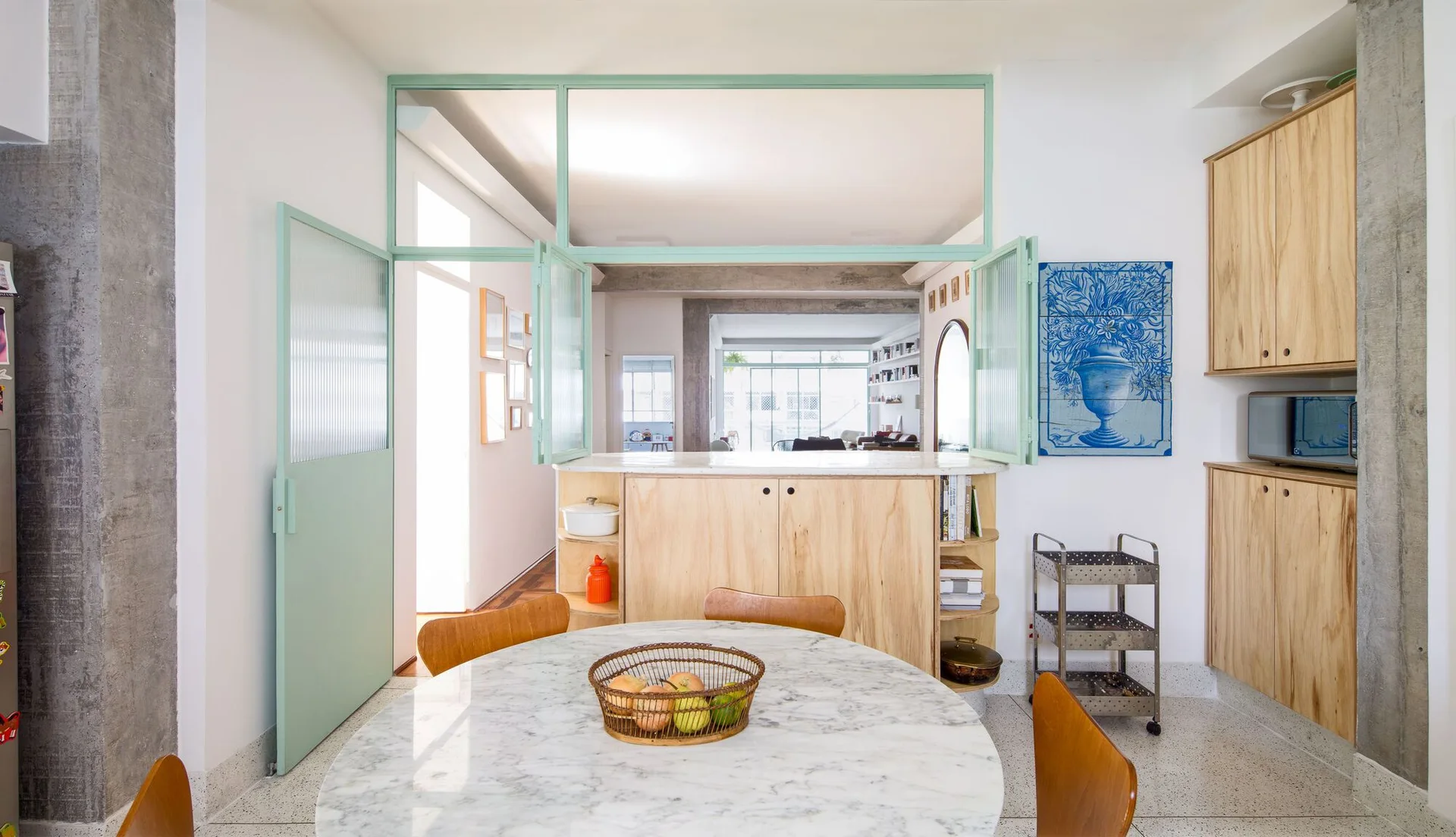 A dining area featuring a round marble table, light wood cabinetry, and a mint green framed glass partition looking into a living space.