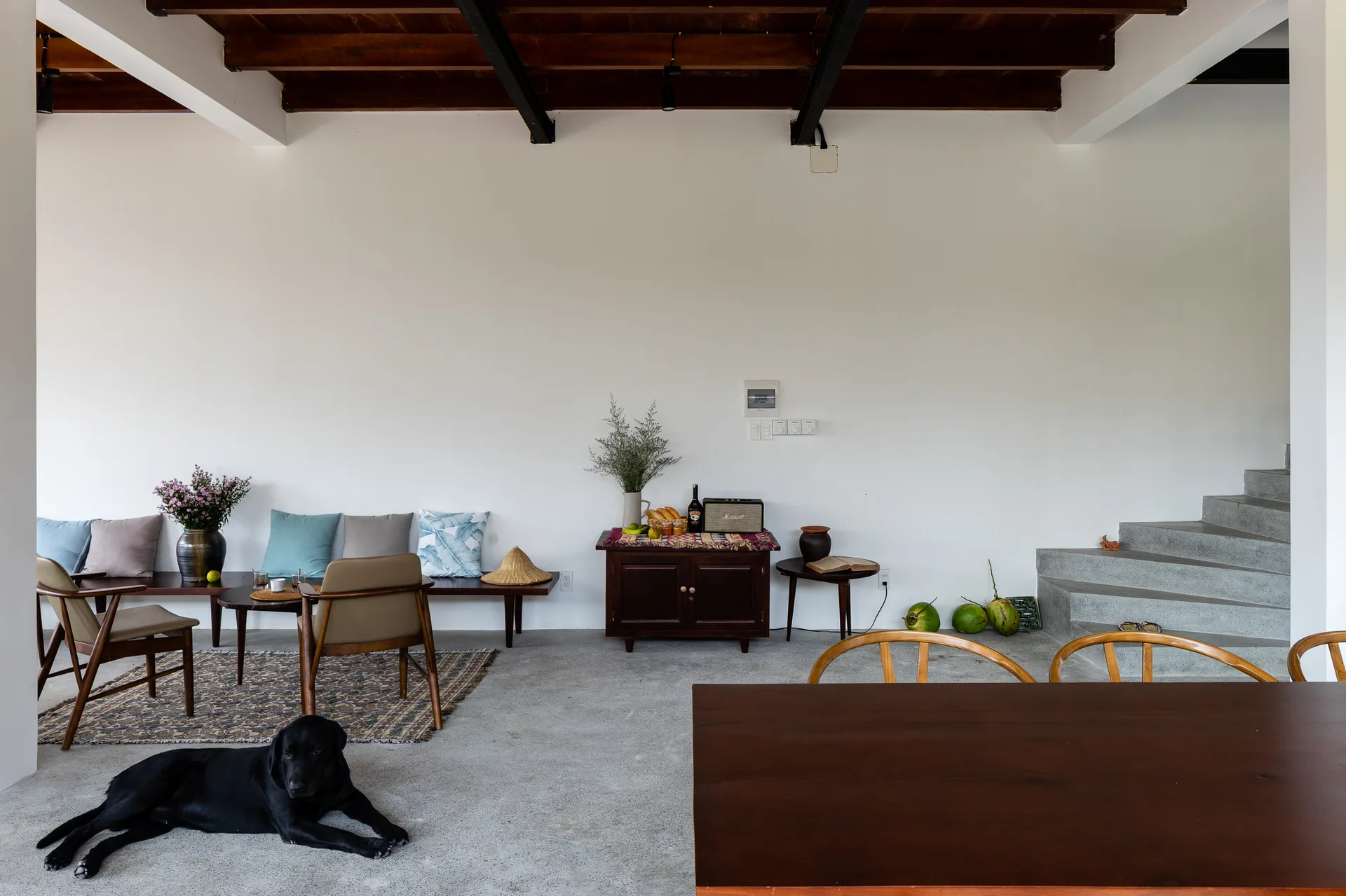 A minimalist open-plan living room features a concrete staircase, exposed dark wood ceiling beams, and low-profile wooden furniture on concrete floors.