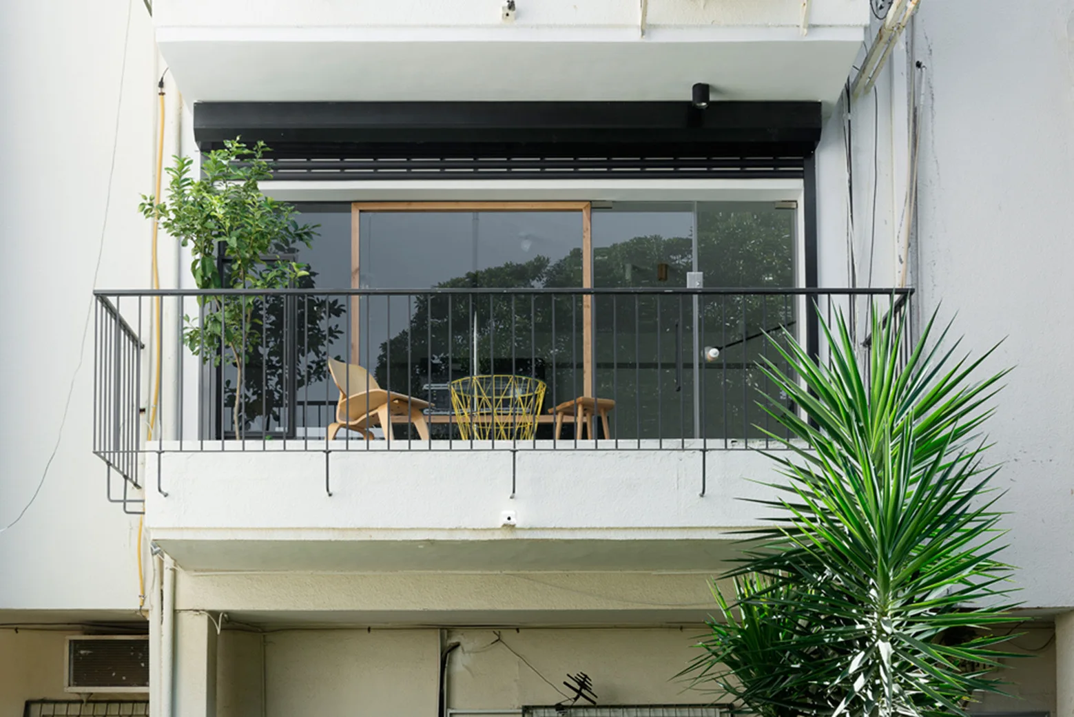 A small apartment balcony with a black metal railing, a wooden chair, a yellow wire side table, and a potted plant.