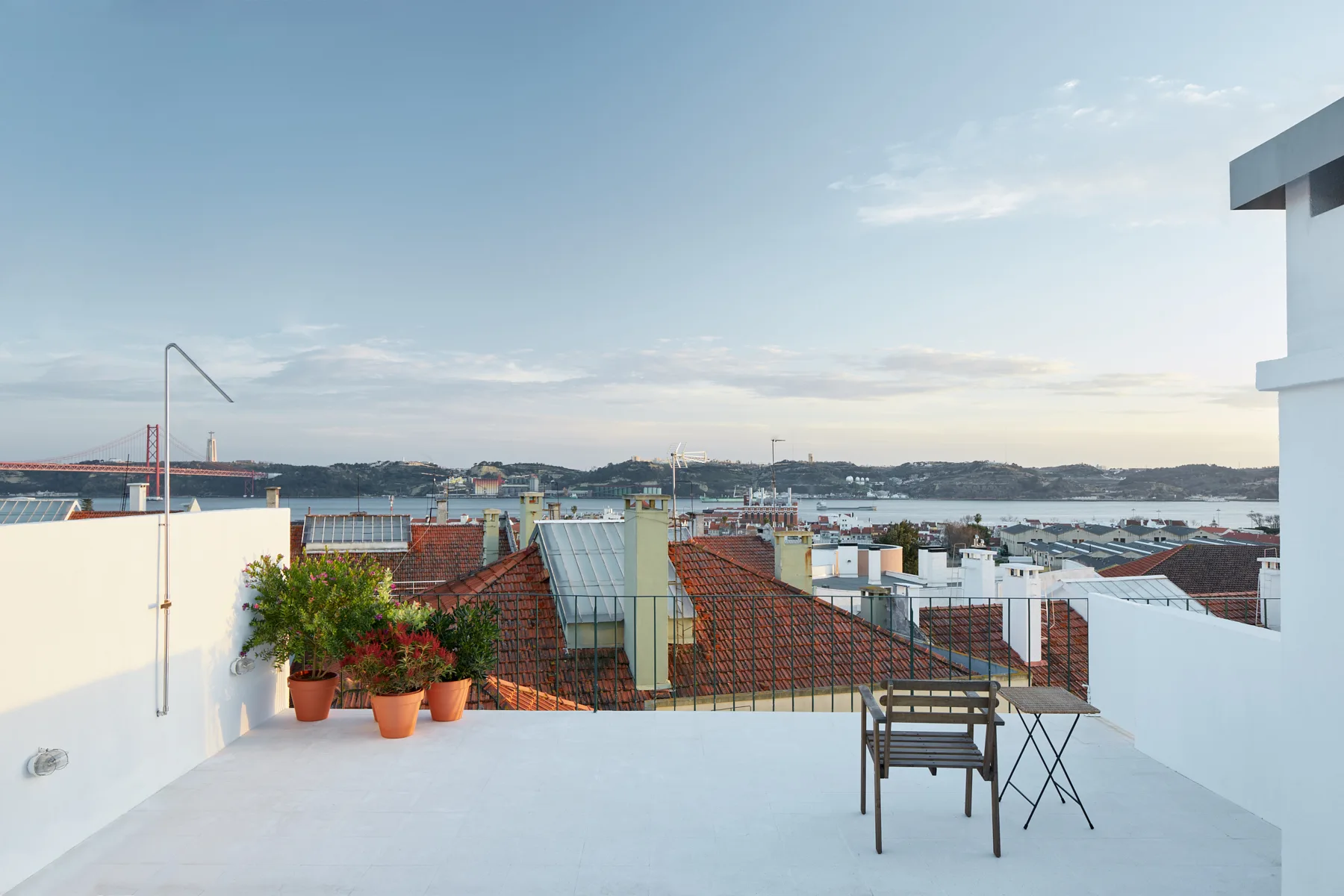 A white rooftop terrace features terracotta potted plants, a wooden chair, a small table, and a view of tiled roofs and a distant bridge.