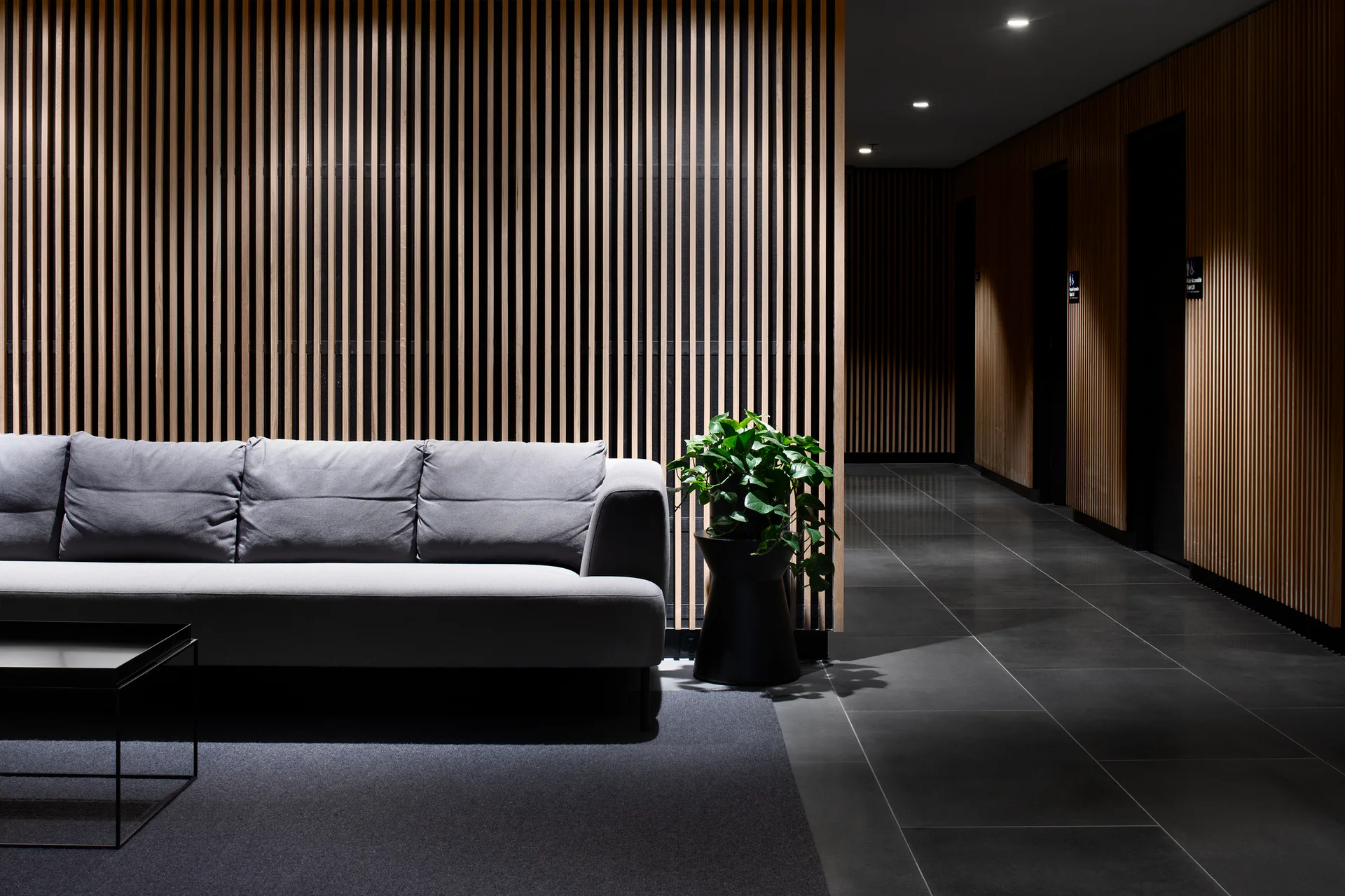 A grey sofa sits against a vertical wood slat wall next to a potted green plant and dark tile flooring in a modern lounge.