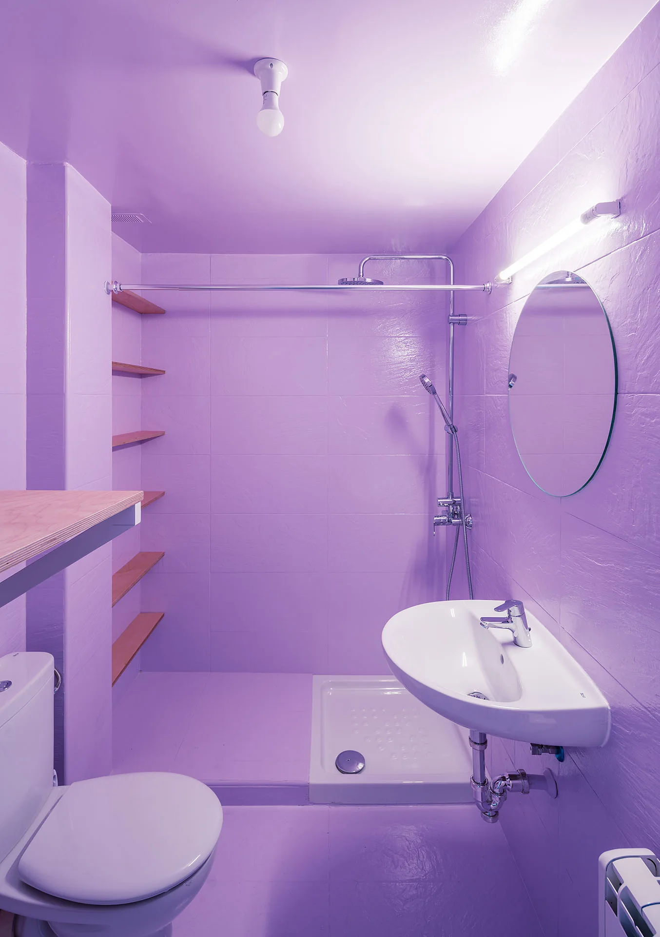 A small bathroom lined in glossy lavender tiles with a wall-mounted sink, a circular mirror, and built-in wood shelving.