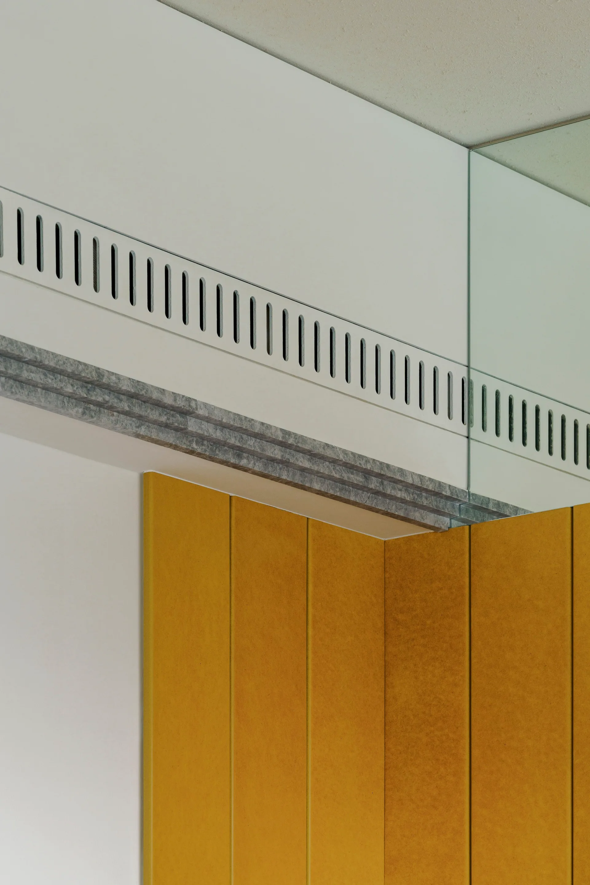 A close-up of vertical mustard yellow wall panels meeting a white soffit with an integrated linear ventilation grille and grey trim.