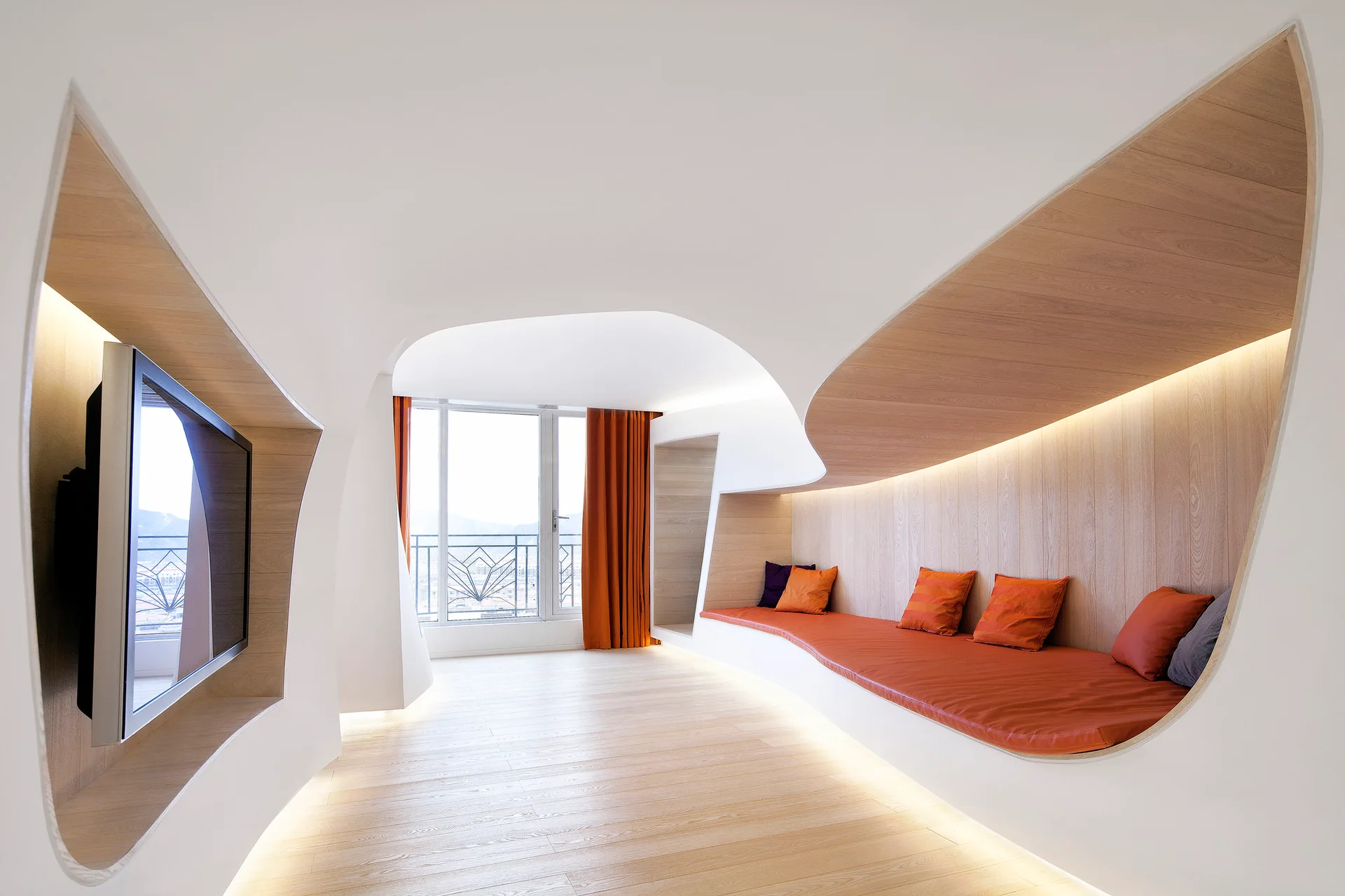 A modern living room features futuristic curved white walls containing a built-in wooden bench with orange cushions and a wall-mounted TV.