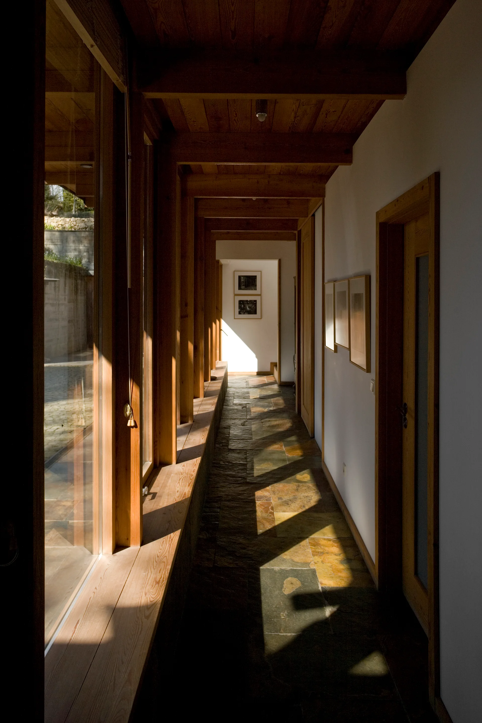 A long interior hallway features exposed wood ceiling beams, textured stone tile flooring, and a floor-to-ceiling glass wall letting in sunlight.
