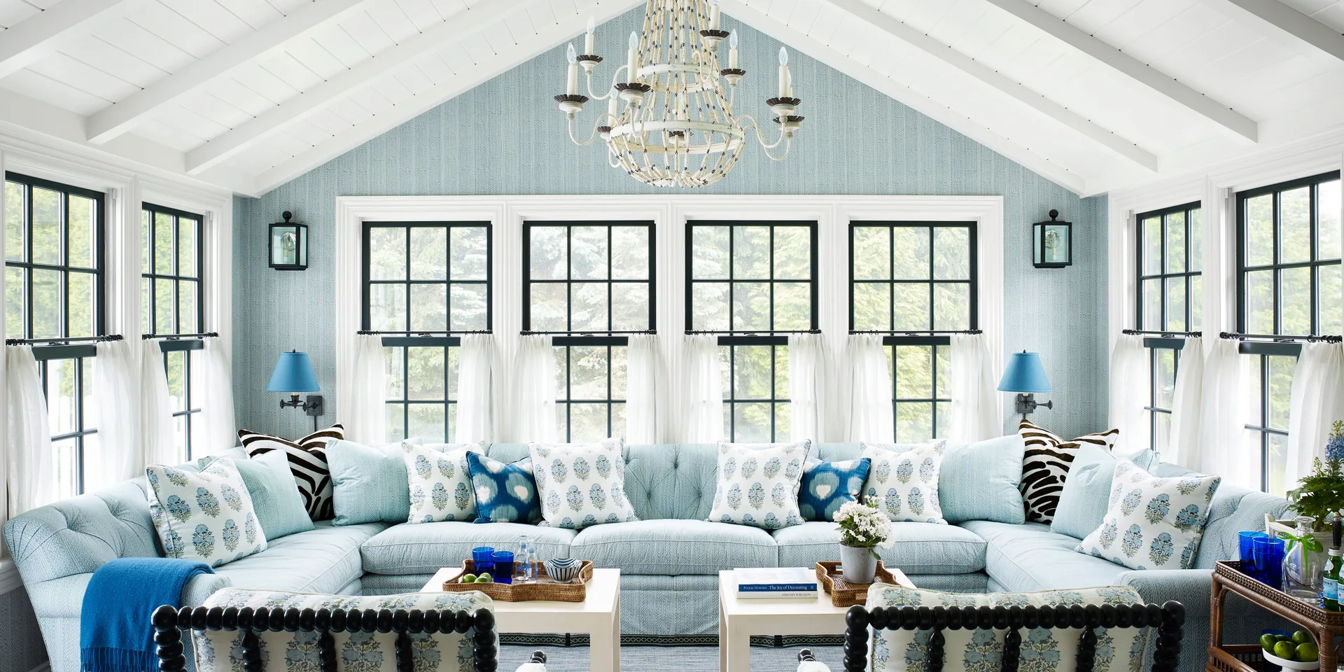 A light blue tufted sectional sofa sits in a bright sunroom beneath a vaulted white ceiling with a beaded chandelier and large black-framed windows.