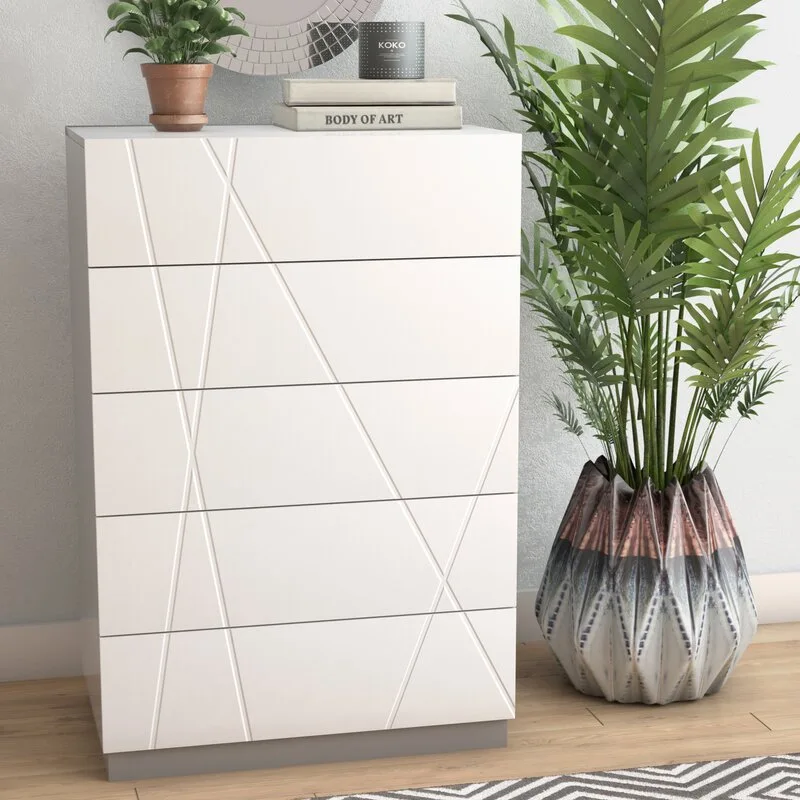 A modern white five-drawer dresser with etched geometric lines on its front stands next to a large potted palm on a light wood floor.