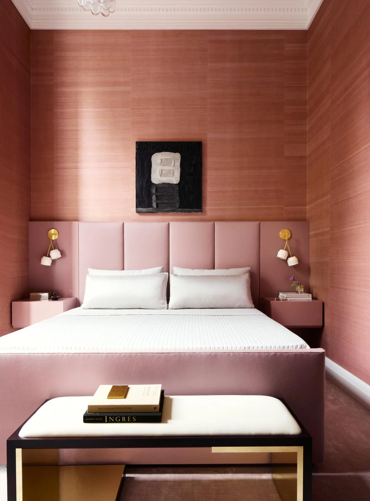 A symmetrical pink bedroom features a mauve upholstered headboard, matching floating nightstands, and brass wall sconces against grasscloth wallpaper.