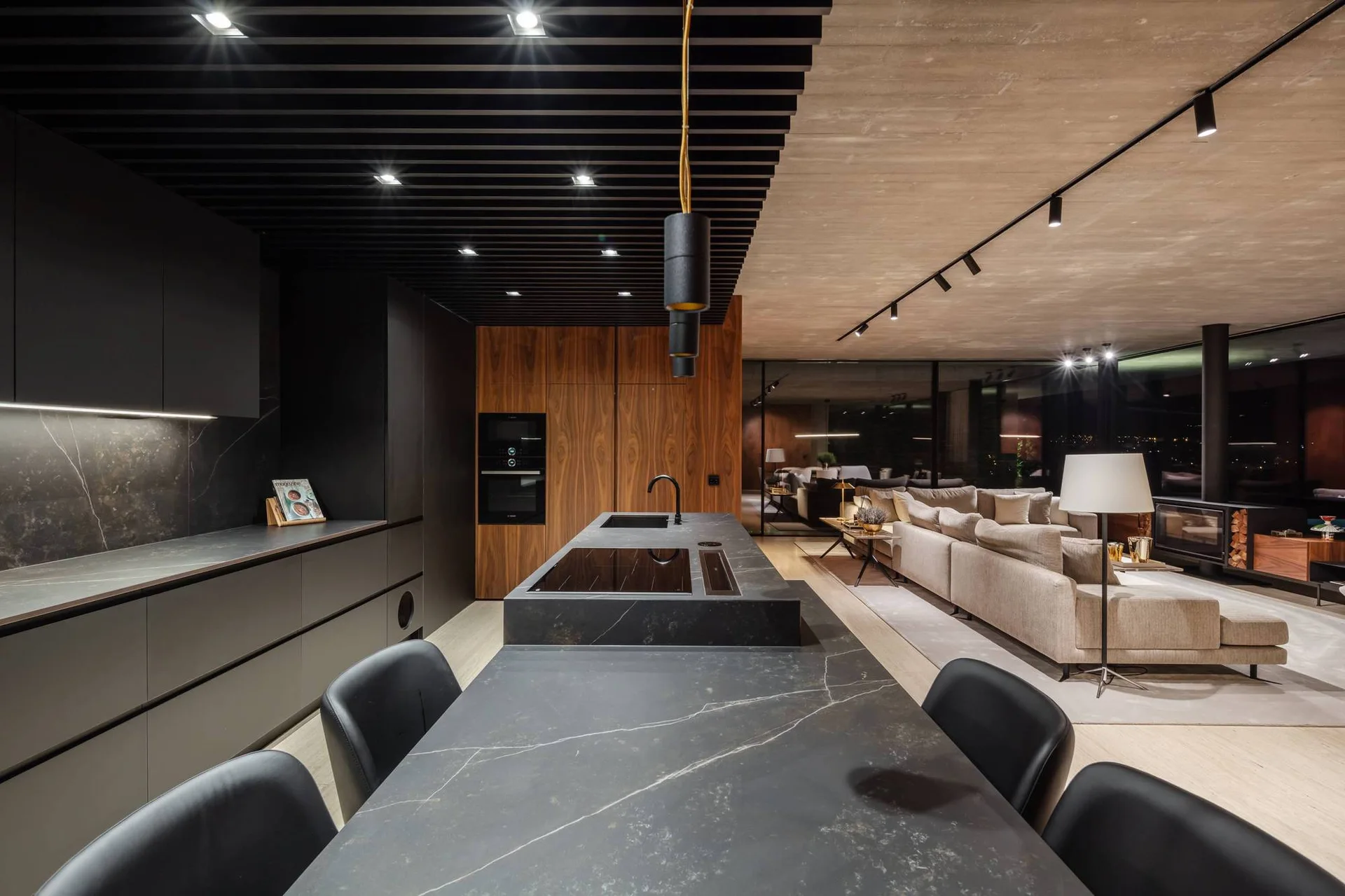 A modern open-plan kitchen features a dark veined stone island, black slatted ceiling, and wood cabinets next to a living area with a beige sofa.