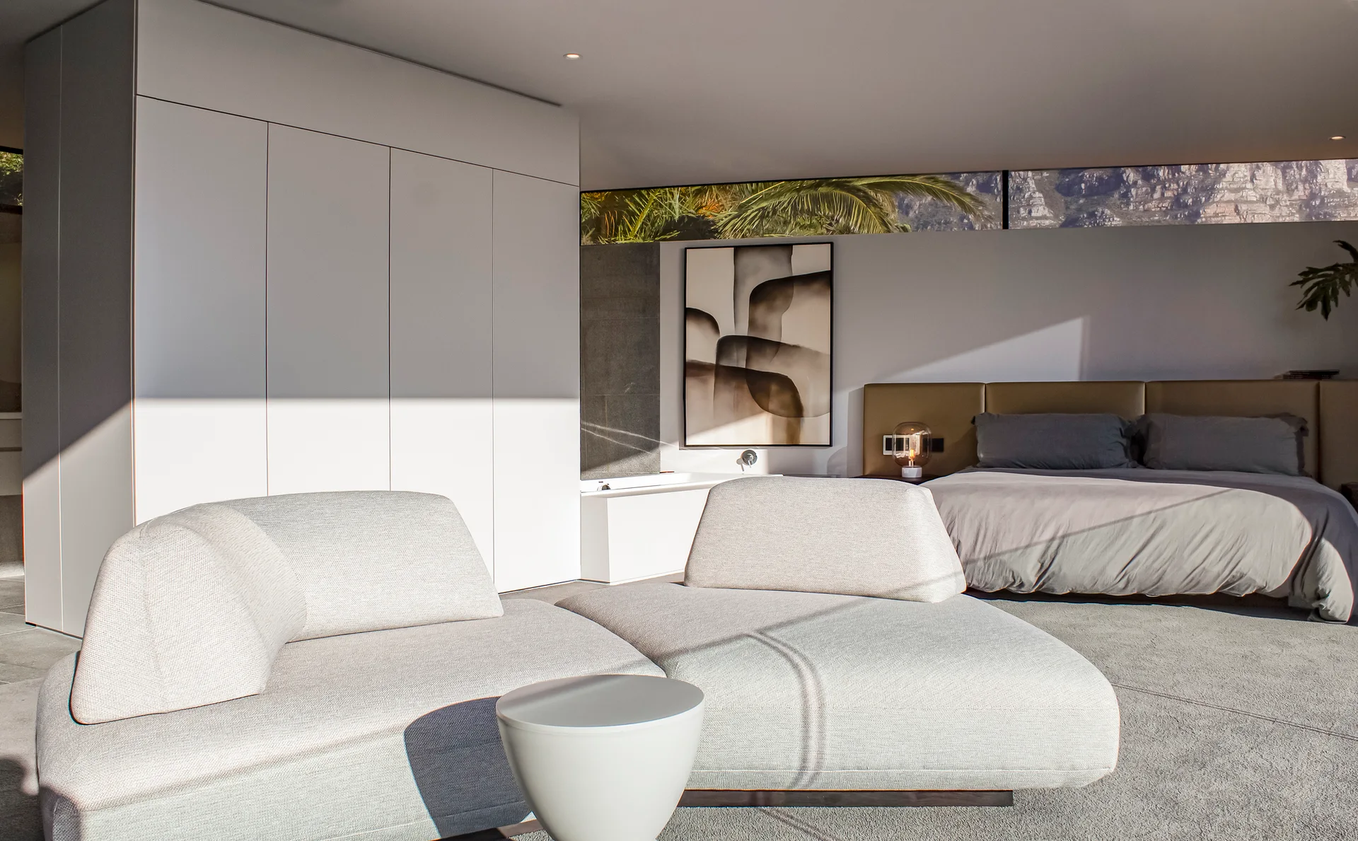 A modern bedroom features a light grey modular sofa in the foreground, a low-profile bed with a tan headboard, and a long clerestory window.