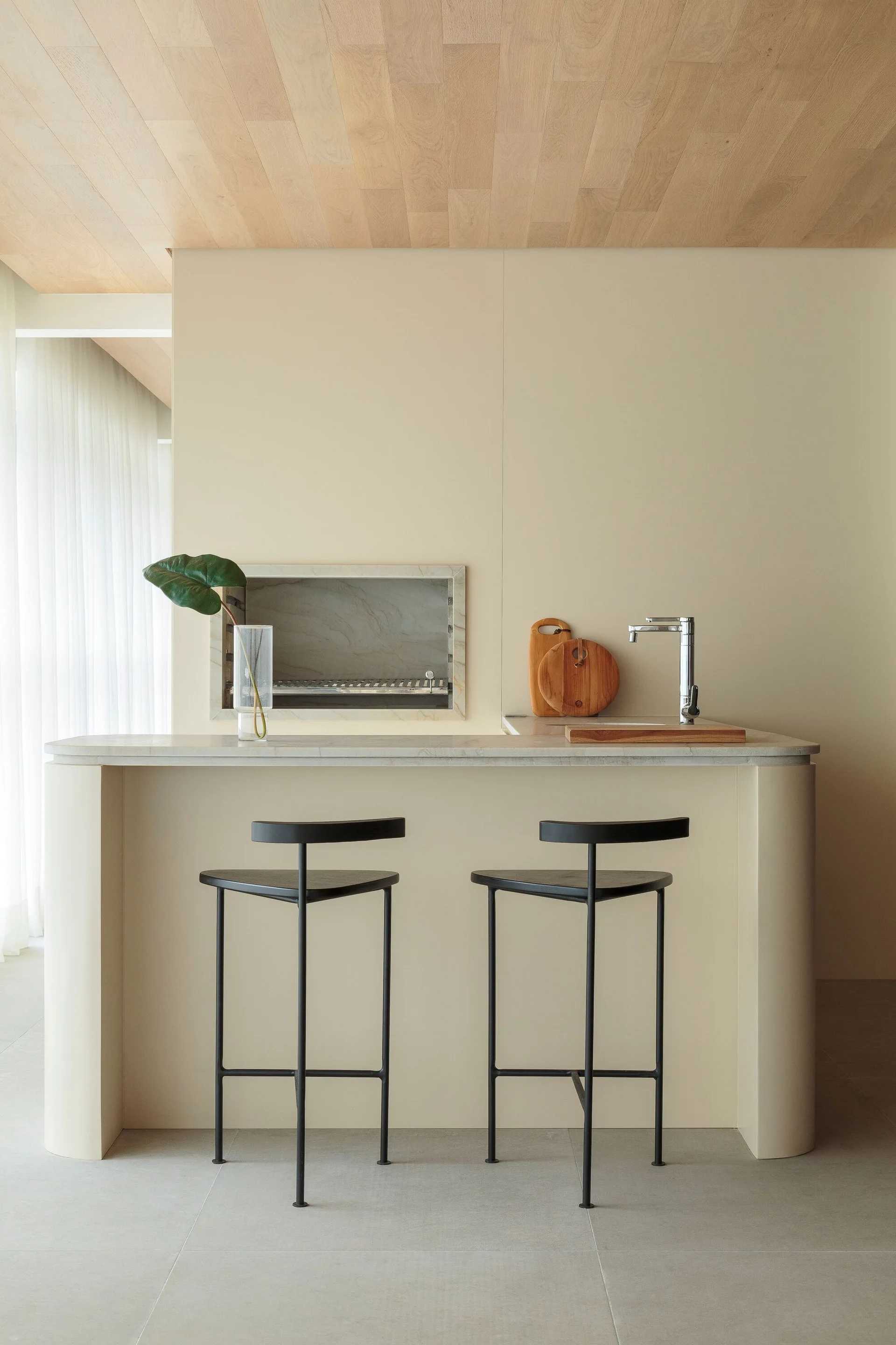 A minimalist beige kitchen island with a curved base and a stone countertop is paired with two slim black bar stools under a light wood ceiling.