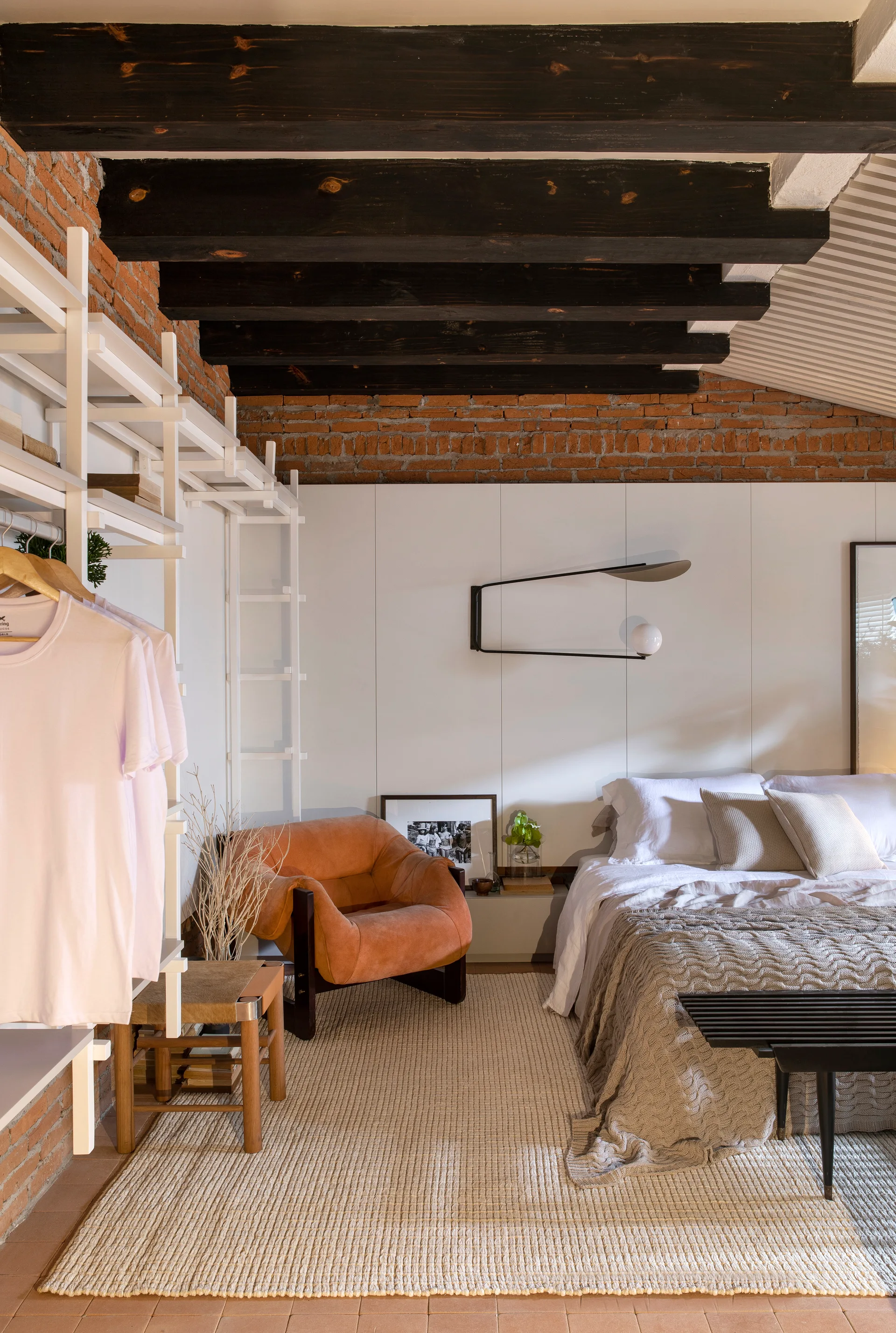 A modern bedroom features a bed with textured linens, a tan leather armchair, white wall paneling, and an exposed brick wall with dark wood beams.