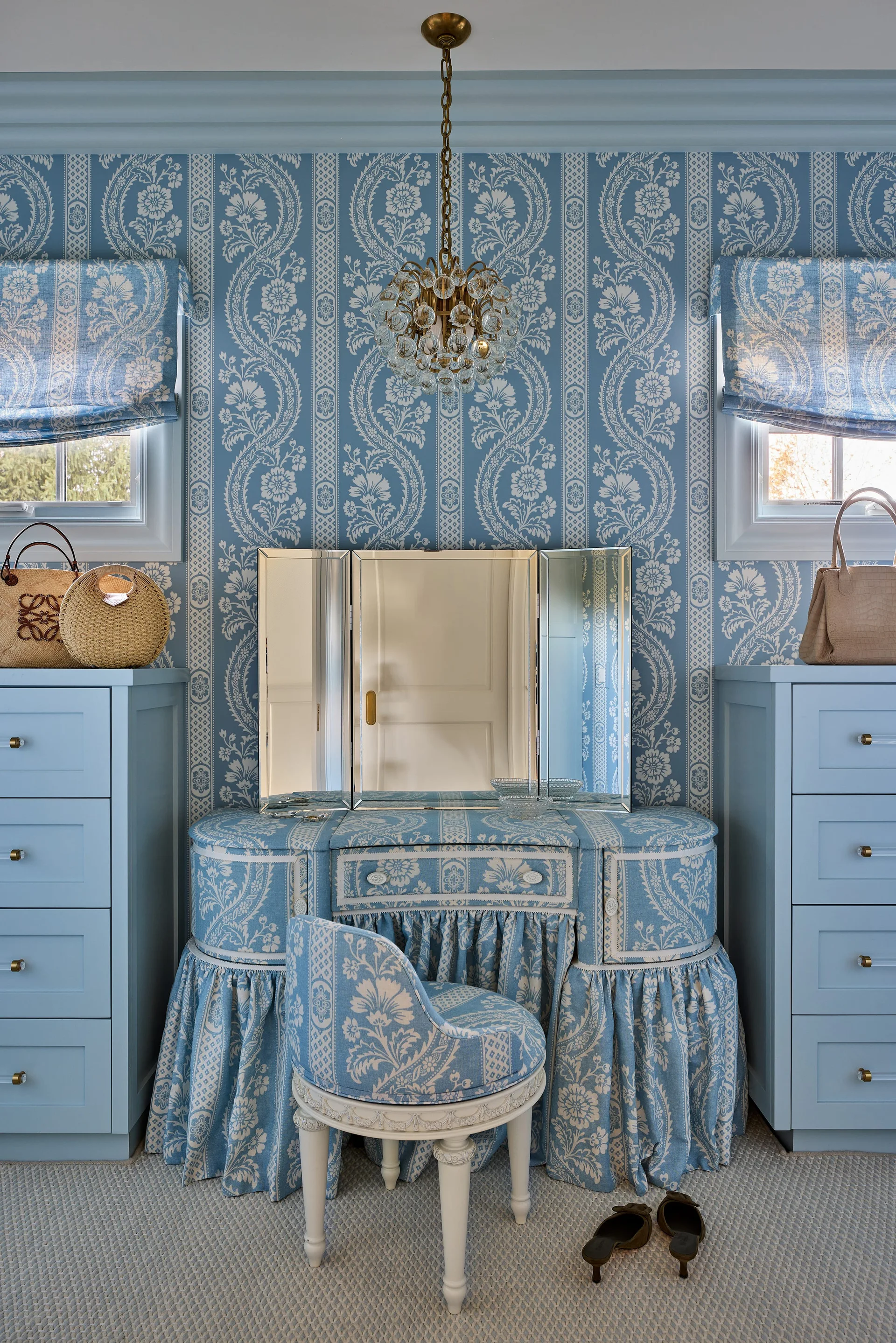 A dressing room vanity area features matching blue floral wallpaper, a skirted vanity table, a tri-fold mirror, and flanking blue dressers.
