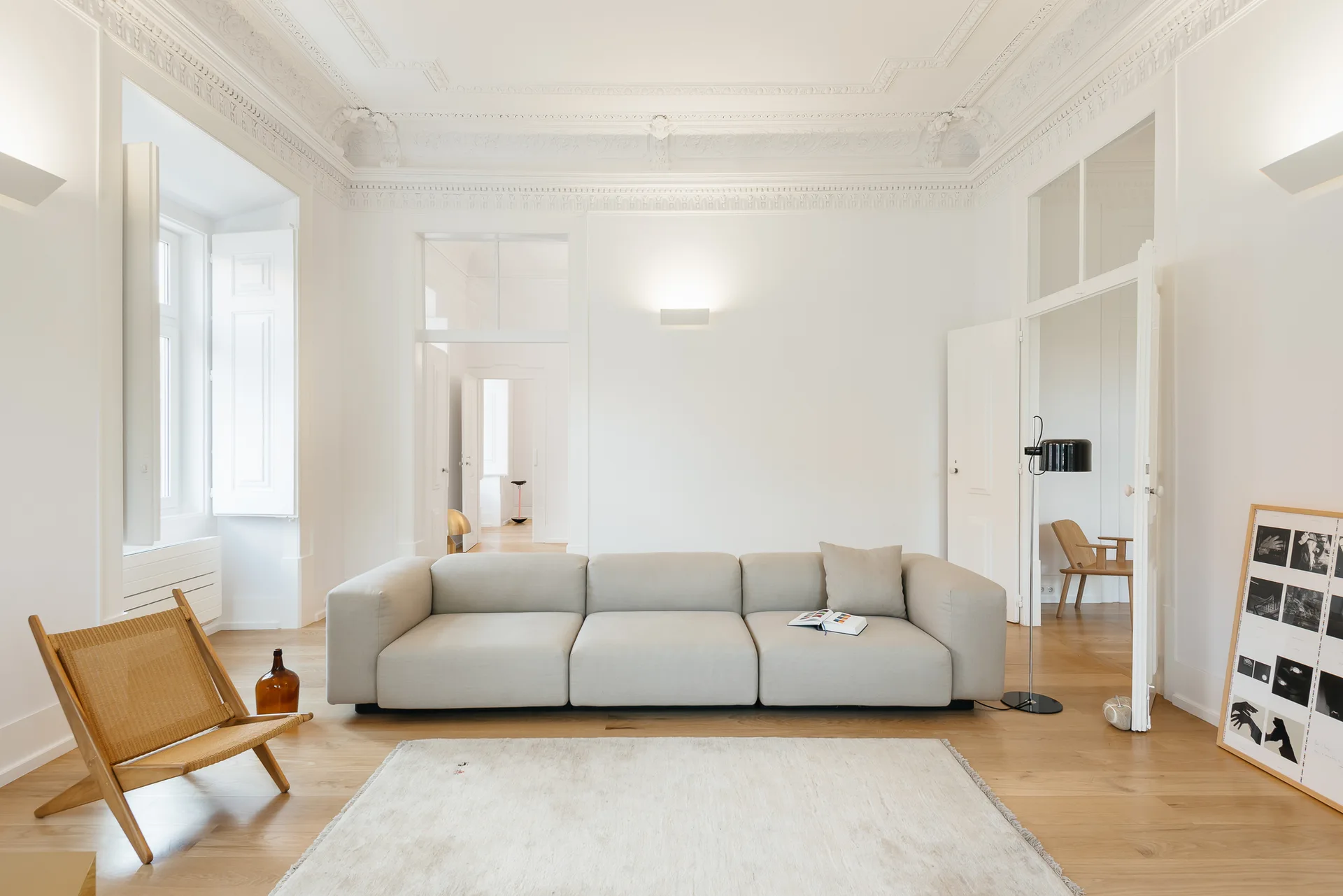 A low-profile beige sofa and a wooden wicker chair sit on a light hardwood floor in a white room with ornate ceiling molding.