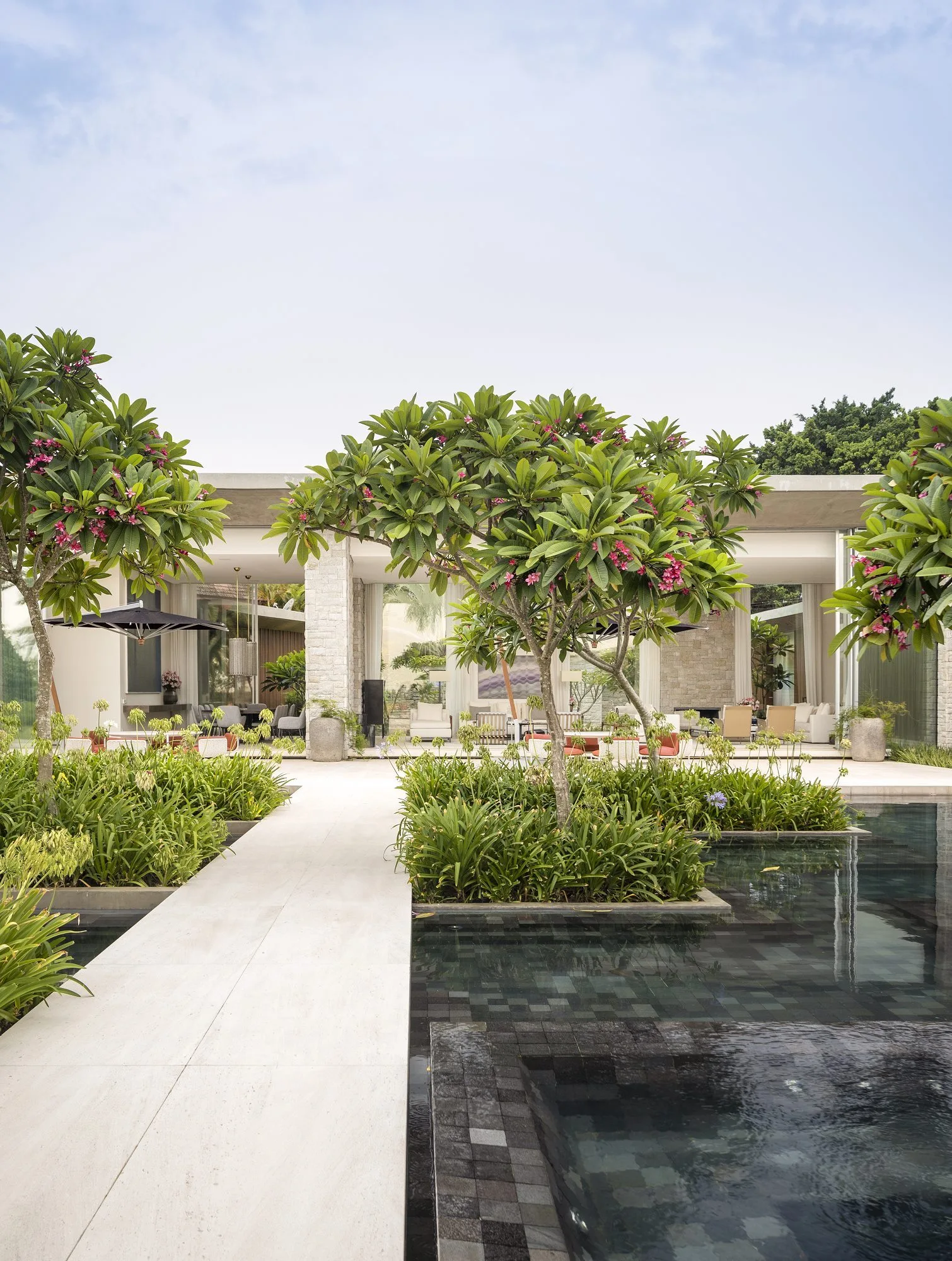 A light stone walkway cuts through a dark tiled pool leading to an open terrace with stone columns and lush plumeria trees.
