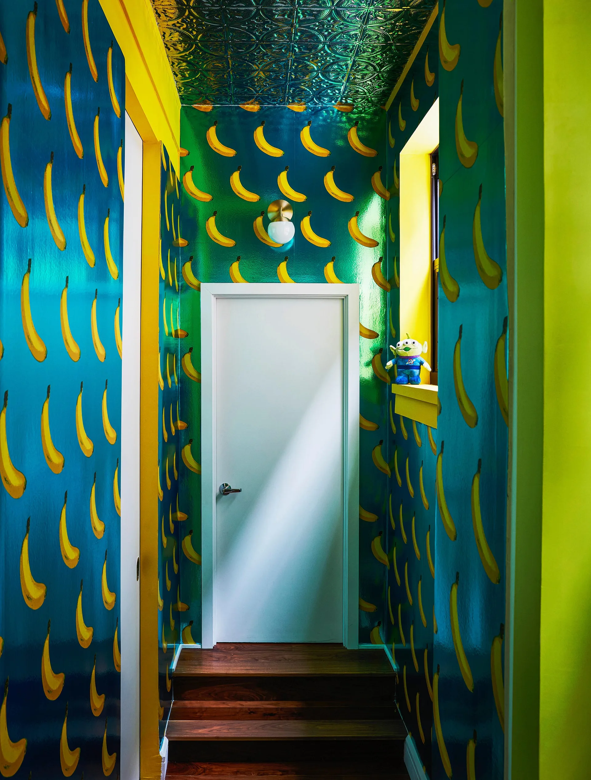 A narrow hallway featuring glossy teal banana-patterned wallpaper, a white door, bright yellow trim, and an embossed metallic ceiling.