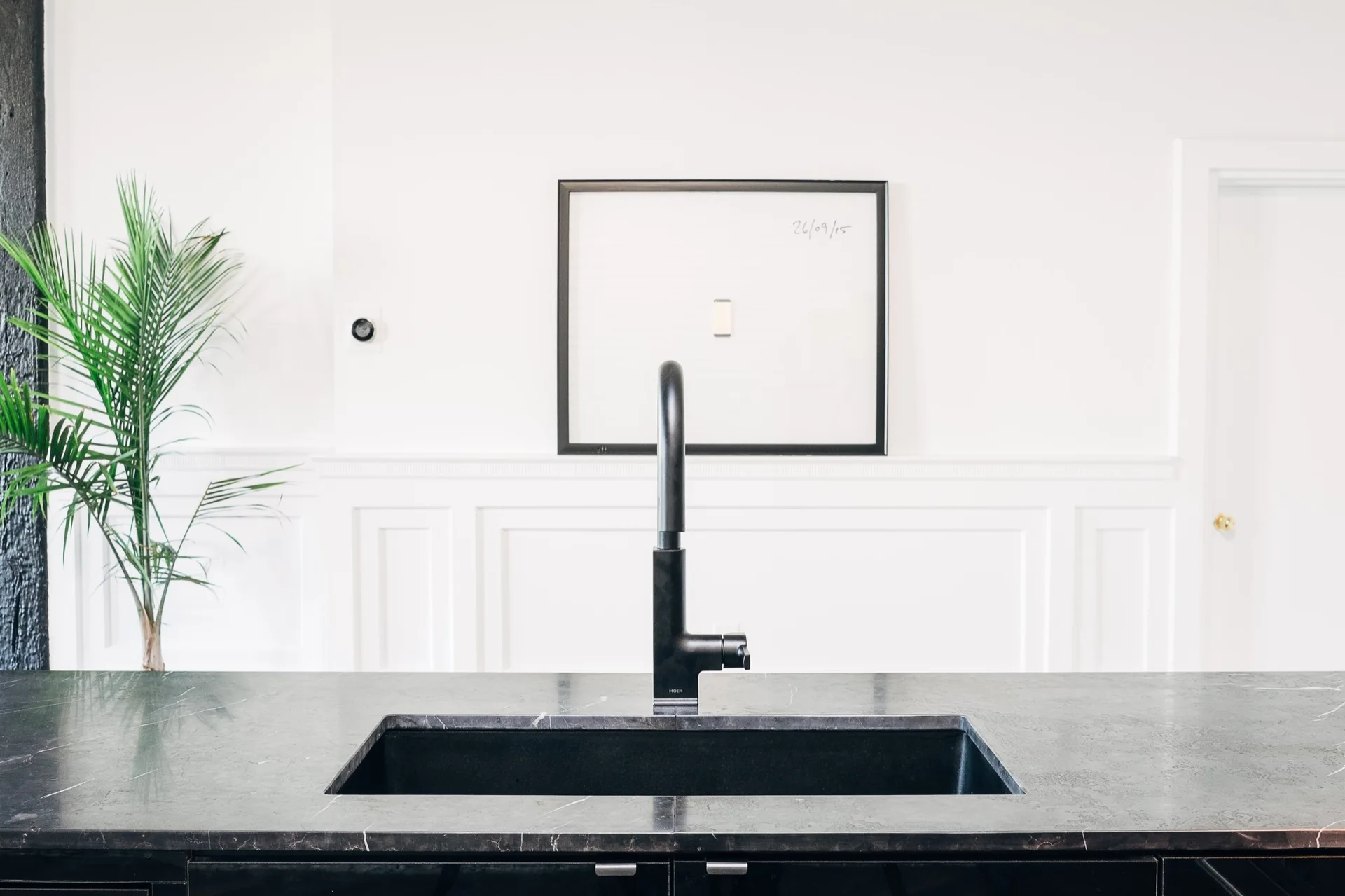 A black matte gooseneck faucet and integrated undermount sink set in a dark veined countertop against a white wall with wainscoting.