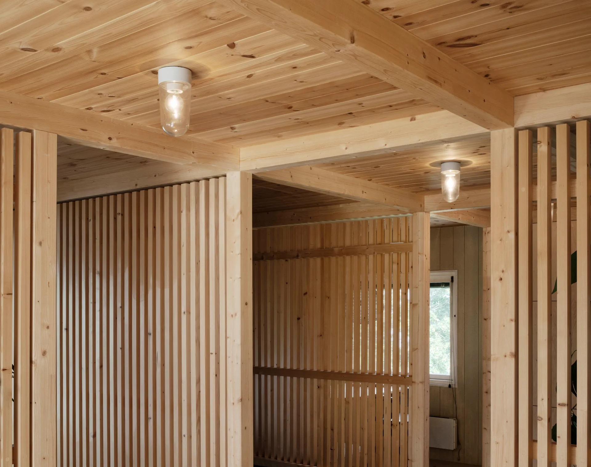 An interior space constructed of light natural wood, featuring vertical slat wall partitions, an exposed beam ceiling, and cylindrical ceiling lights.