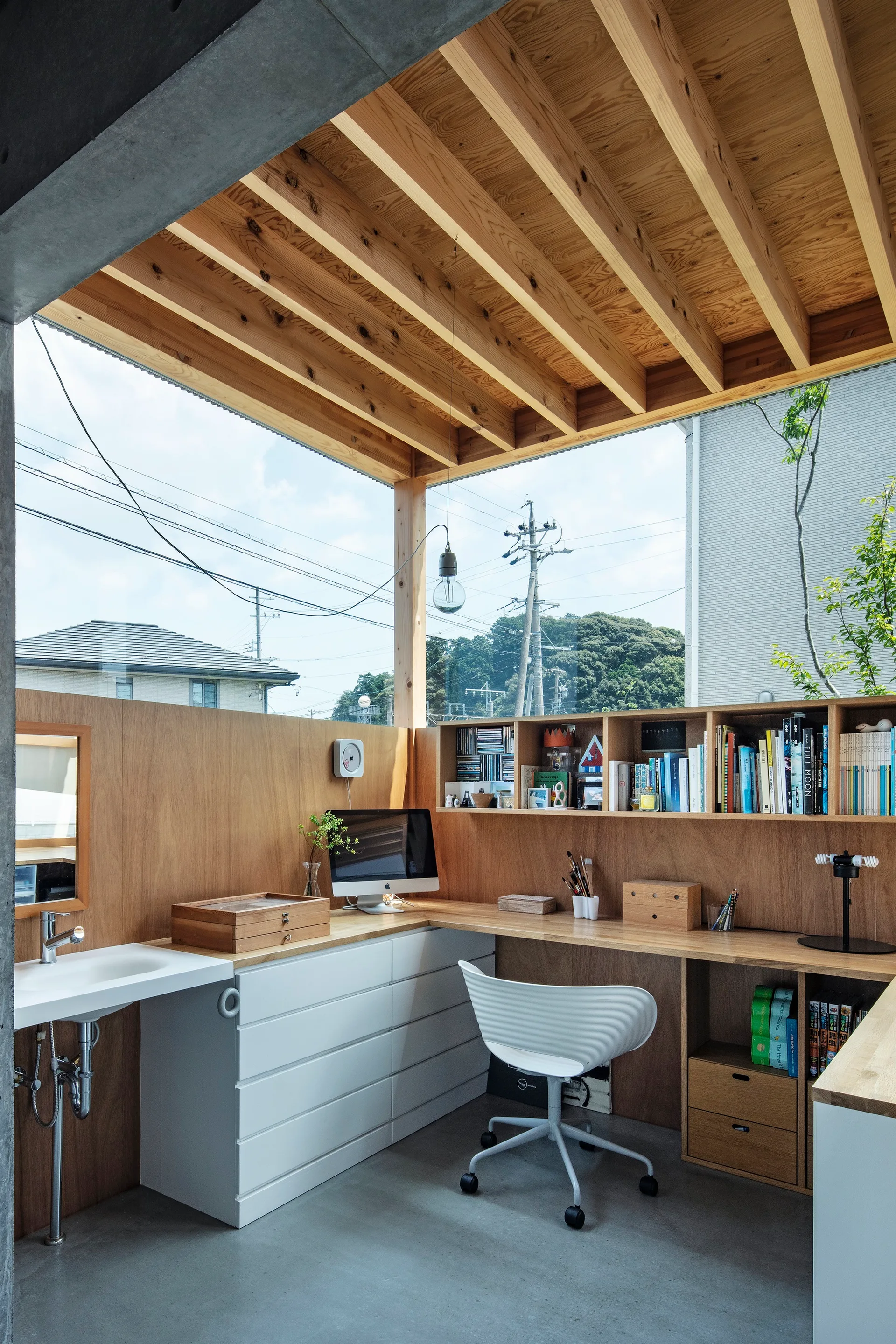 A modern home office features a built-in wooden corner desk, a white swivel chair, a wall-mounted sink, and exposed ceiling beams below a large window.