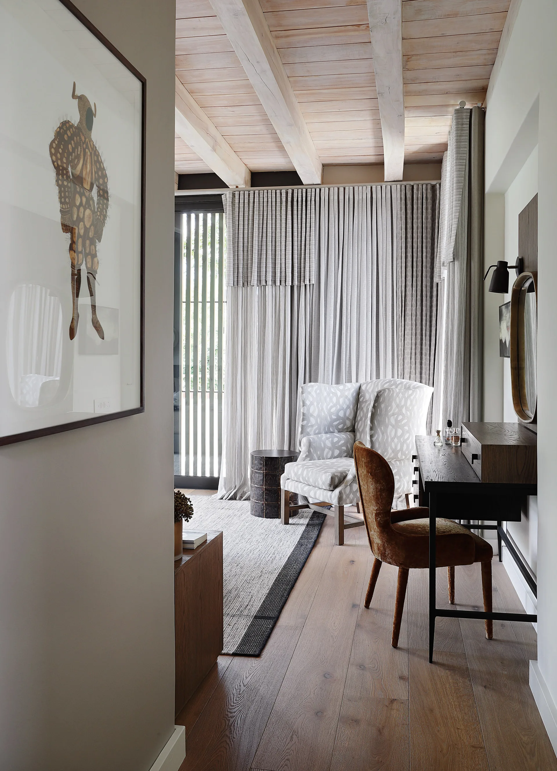 A bedroom alcove shows a dark wood desk with a velvet chair, a patterned armchair, and grey drapes under exposed ceiling beams.