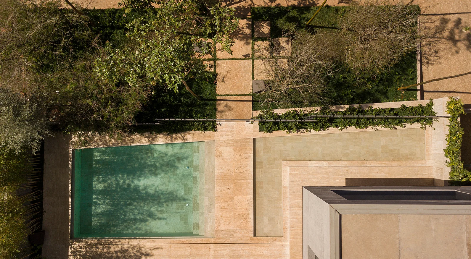 An overhead aerial view of a rectangular swimming pool next to tan stone pavers, green garden plantings, and a stone walkway.