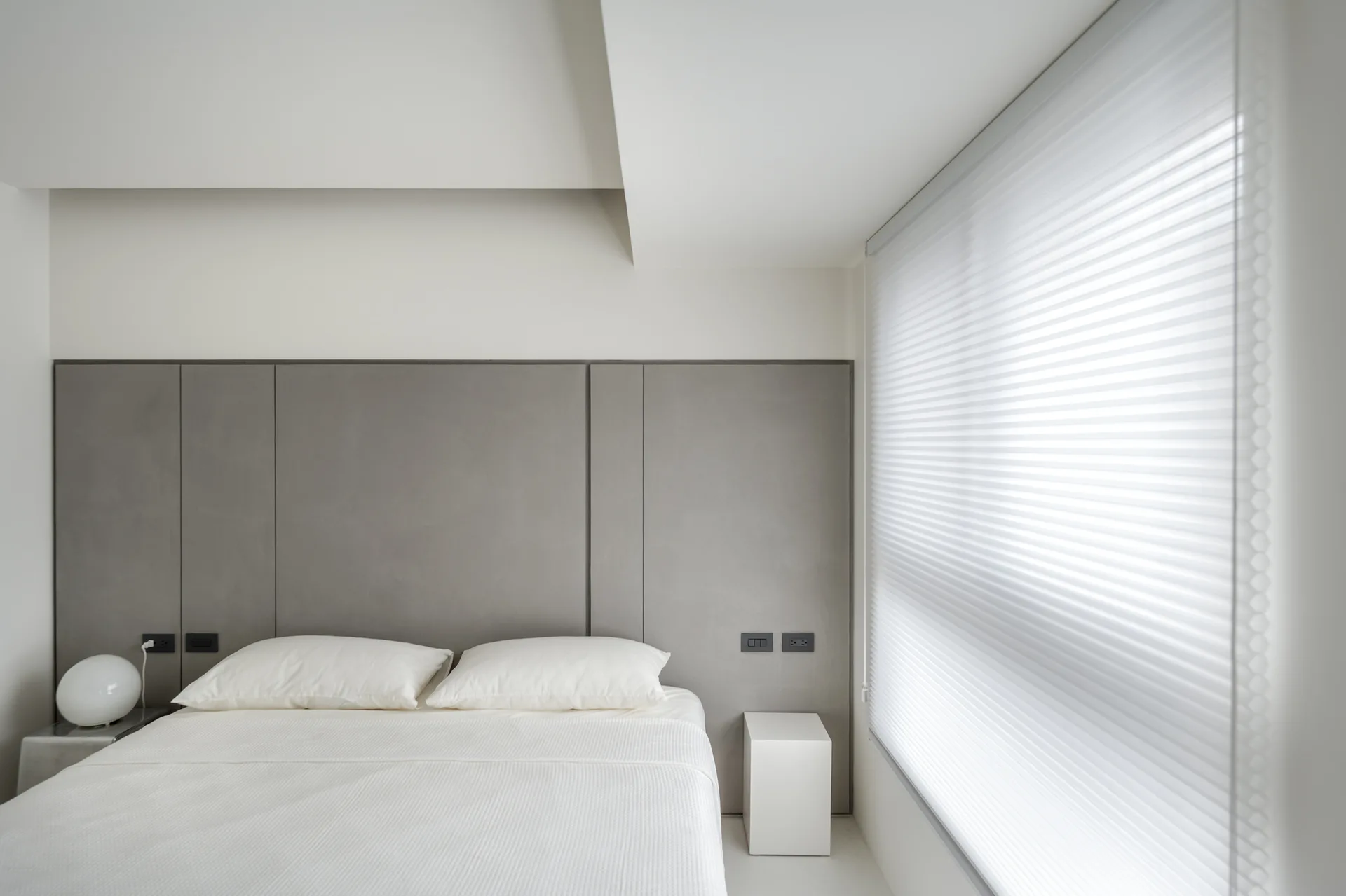 A minimalist bedroom features a bed with white bedding set against a gray paneled headboard wall next to a window with pleated blinds.
