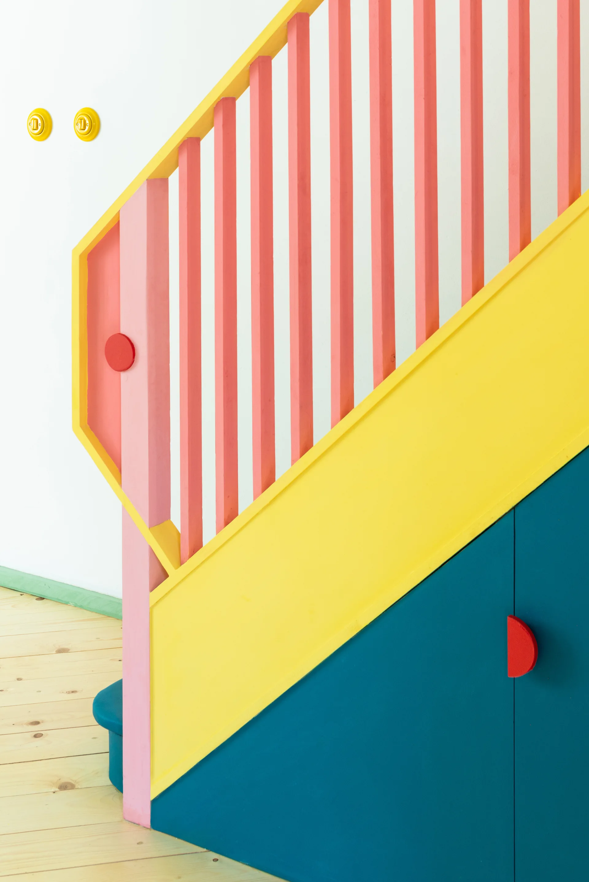 A colorful wooden staircase features a yellow and pink geometric balustrade above teal blue under-stair storage cabinets on a light wood floor.