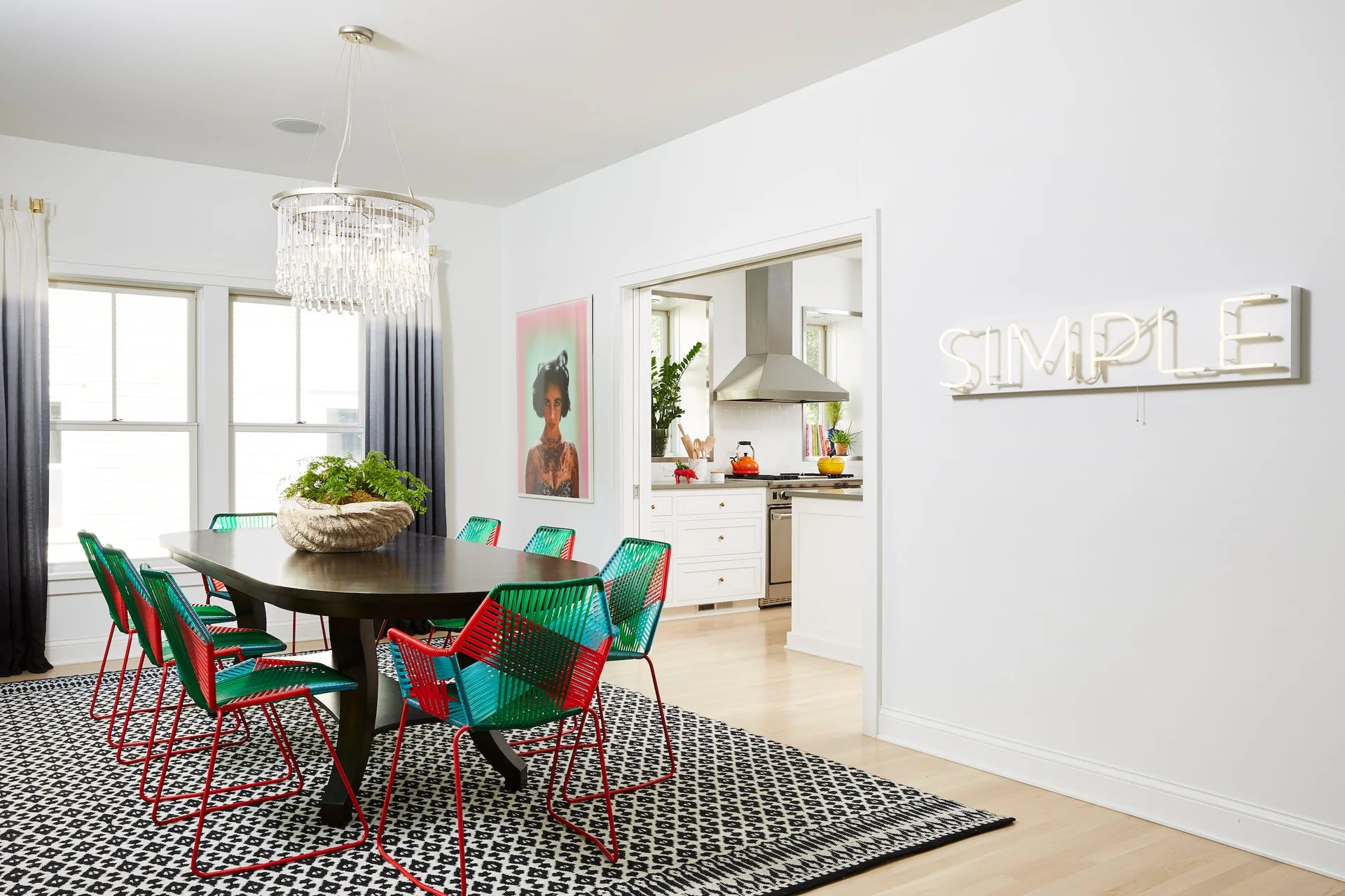 A bright dining room features a dark wood table with colorful woven green and red chairs on a geometric black-and-white patterned rug.