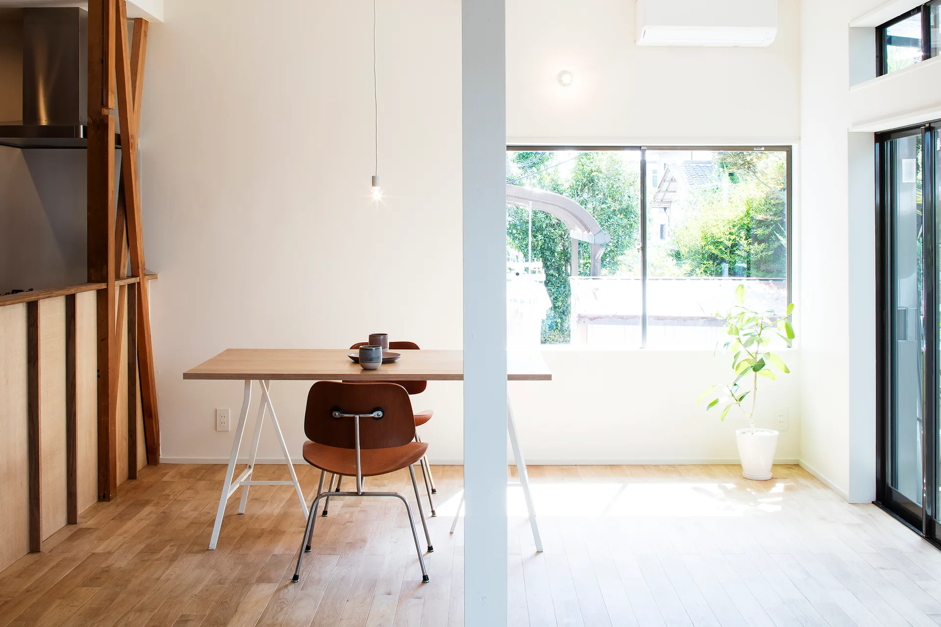 A minimalist dining room featuring a wooden tabletop on white trestle legs, mid-century wooden chairs, and a large window.
