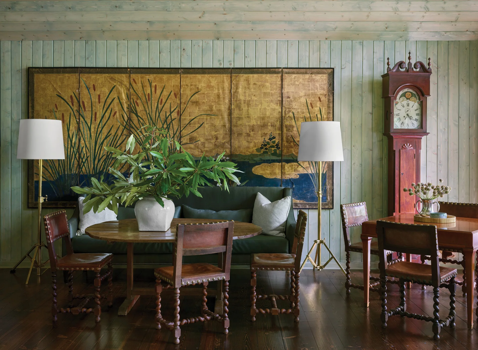 A dining room with light green plank walls features a round table, barley twist chairs, a green sofa, and a large gold screen.