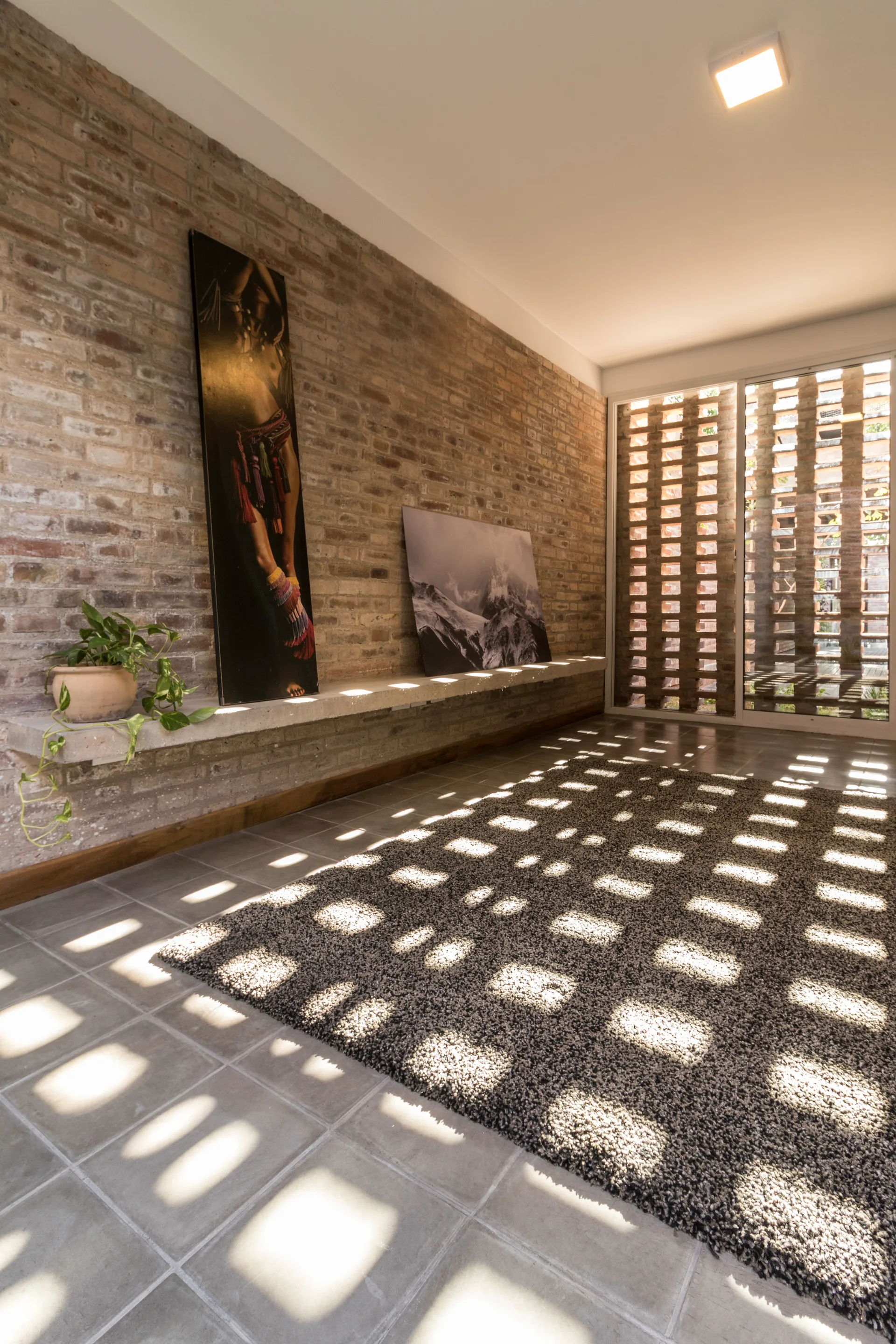 An exposed brick wall features a concrete floating shelf with leaning artwork, while a checkered shadow pattern from a lattice window covers the grey rug.