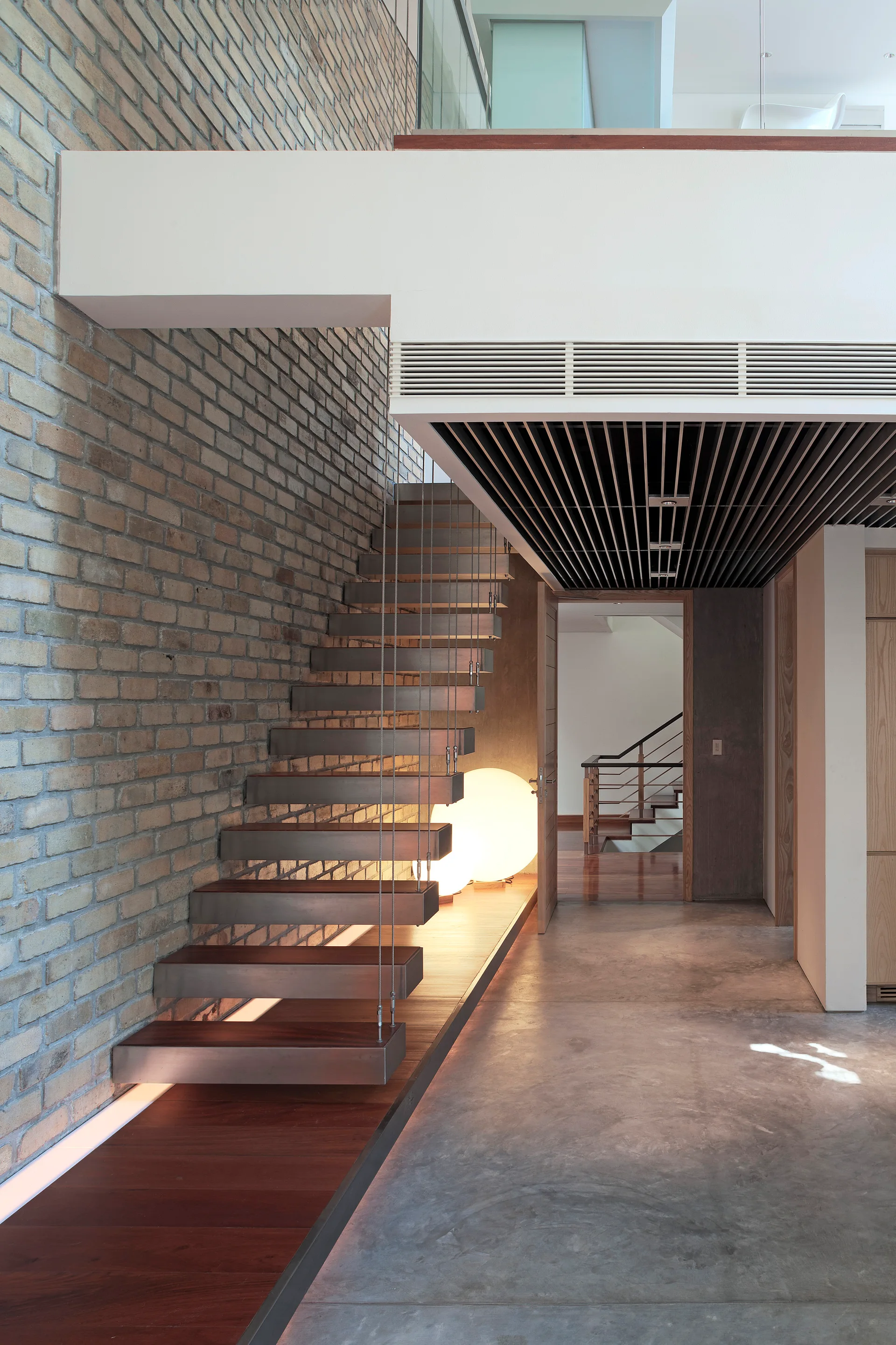 A modern floating staircase with dark wood treads and steel cable supports rises against an exposed brick wall above a polished concrete floor.