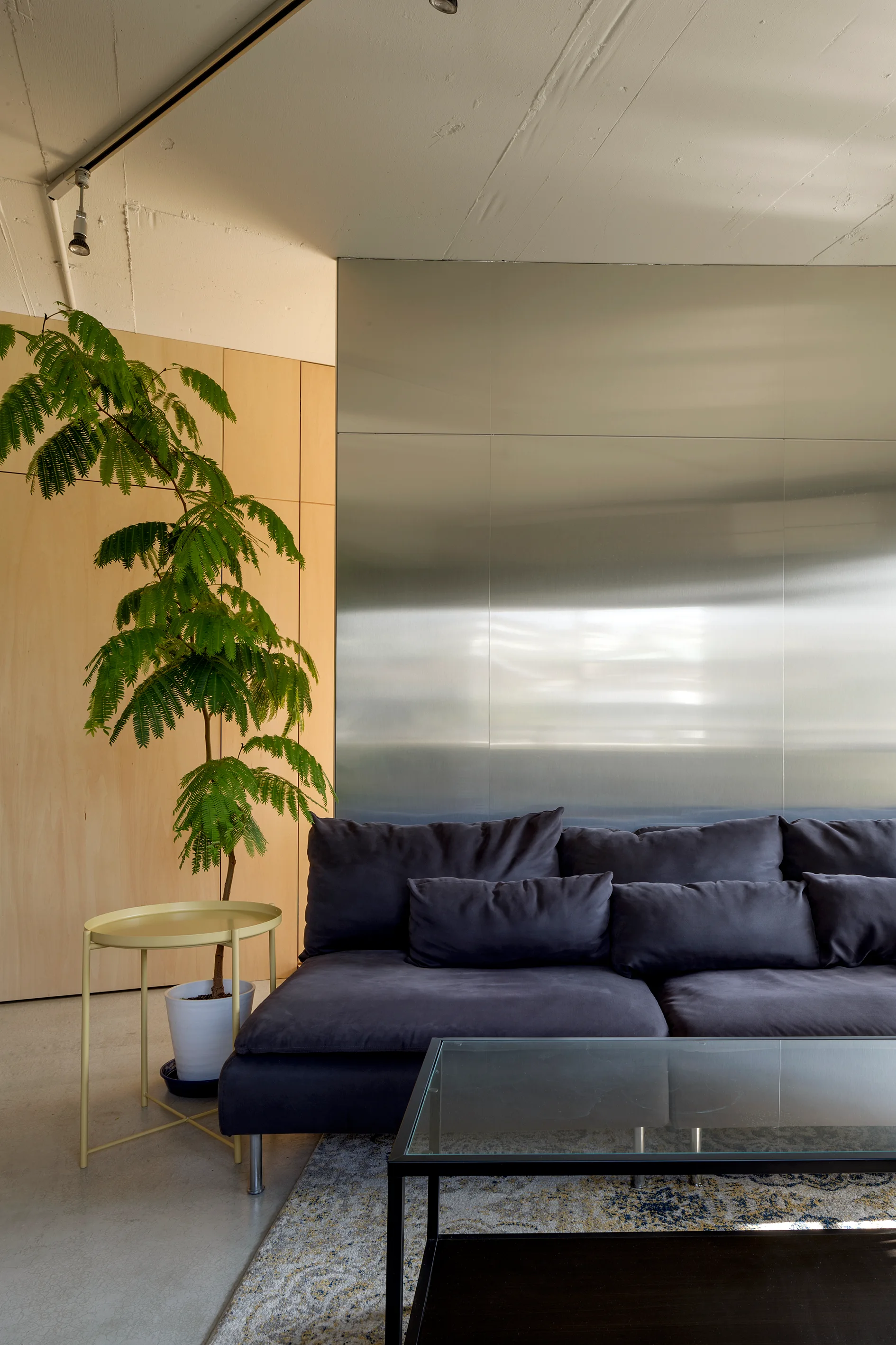 A dark grey sofa sits against a brushed metal wall panel in a living room with light wood paneling, a concrete ceiling, and a yellow side table.