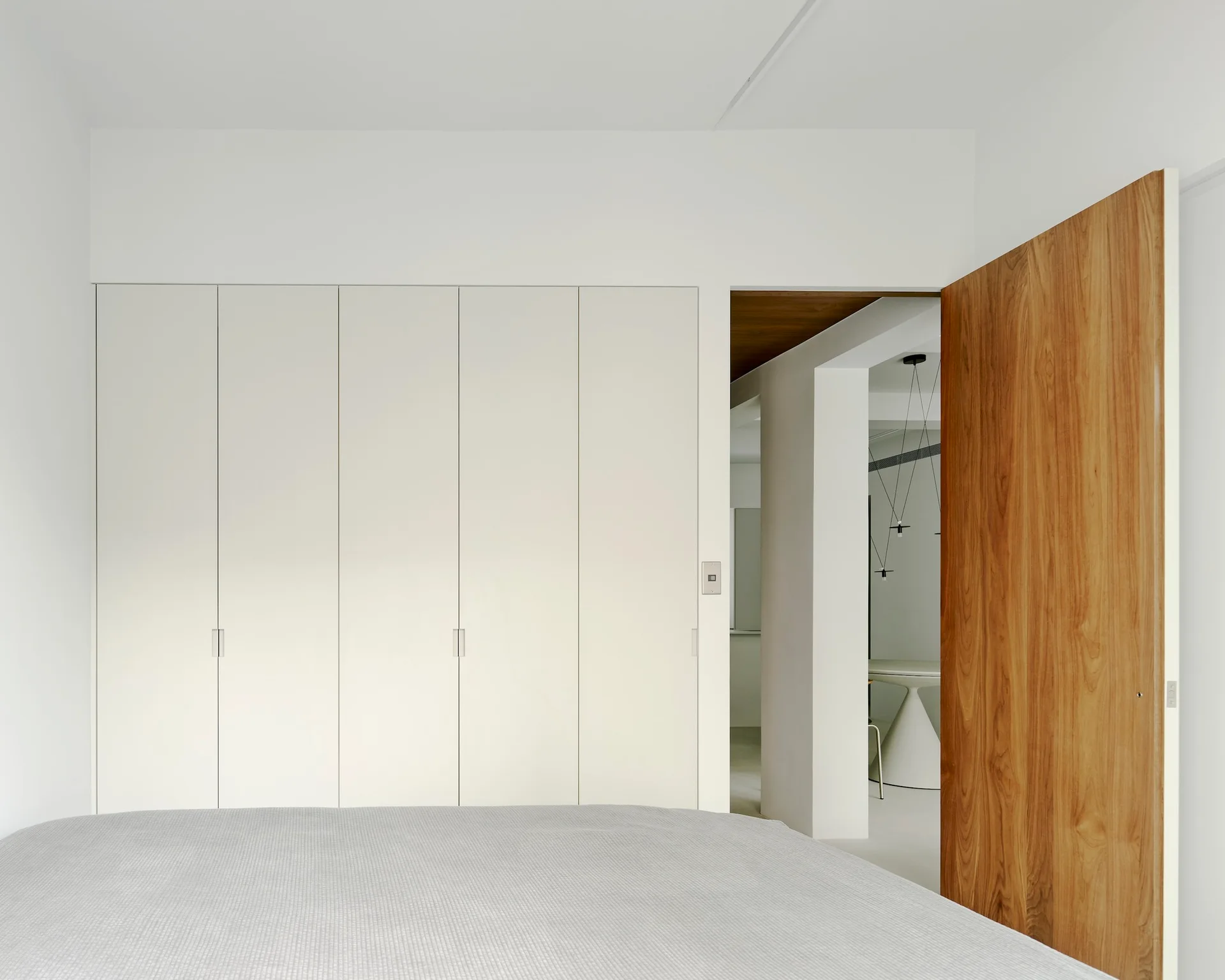 A minimalist white bedroom features a floor-to-ceiling built-in wardrobe and a large open natural wood door leading to an adjacent room.