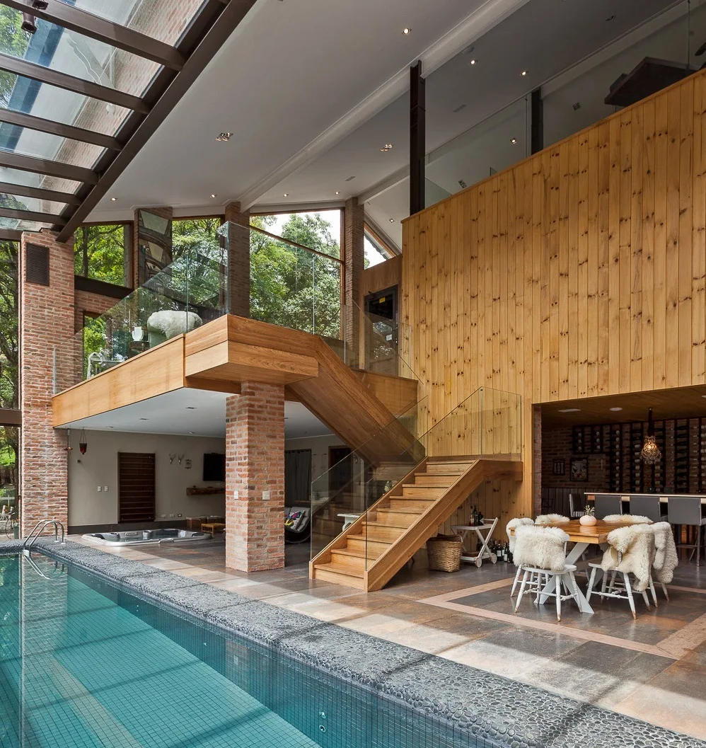 An indoor pool area features a wooden staircase with glass railings, exposed brick columns, and a dining table set under a skylight.