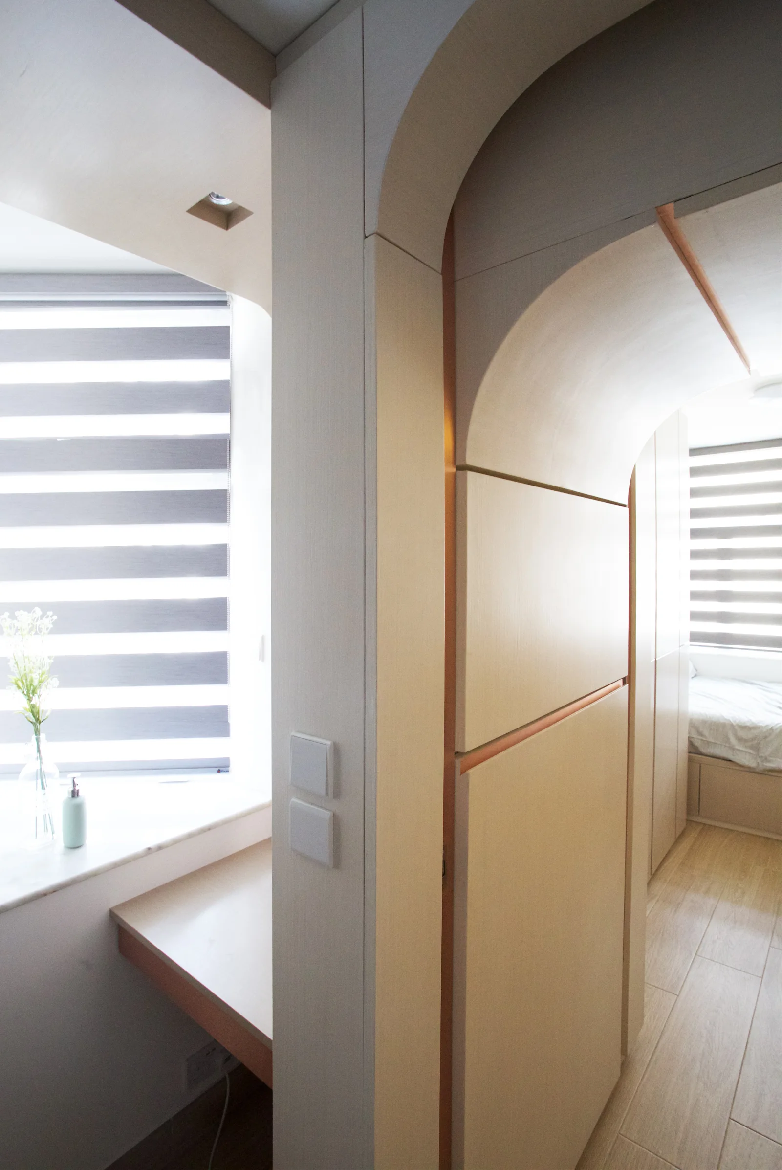 A light wood arched partition separates a small window-side desk from a bedroom featuring striped roller shades and light-colored floors.