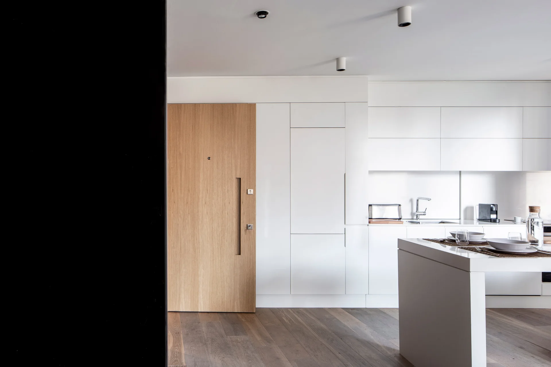 A minimalist white kitchen features handleless cabinets, a flush oak wood door, a white kitchen island with tableware, and dark wood plank floors.